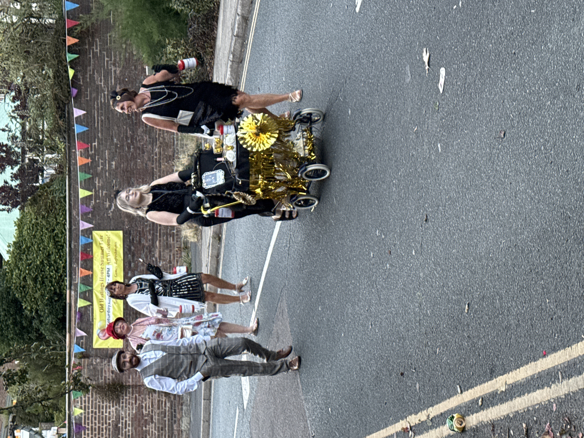 Four people in 1920s attire are walking down a residential street in St Leonards, United Kingdom, during daylight. The street is paved with asphalt and marked with double yellow lines. To the left of the road, a low brick wall is topped with a festive banner. A woman in a black sequined dress and a headdress pushes a decorated pram. Another woman, also in 1920s attire, holds a can. A man in a tweed suit and flat cap walks ahead of them. A fourth person, in a floral dress and a red hat, is partially visible behind them. The background features trees and buildings. The overall scene suggests a community event or celebration.
