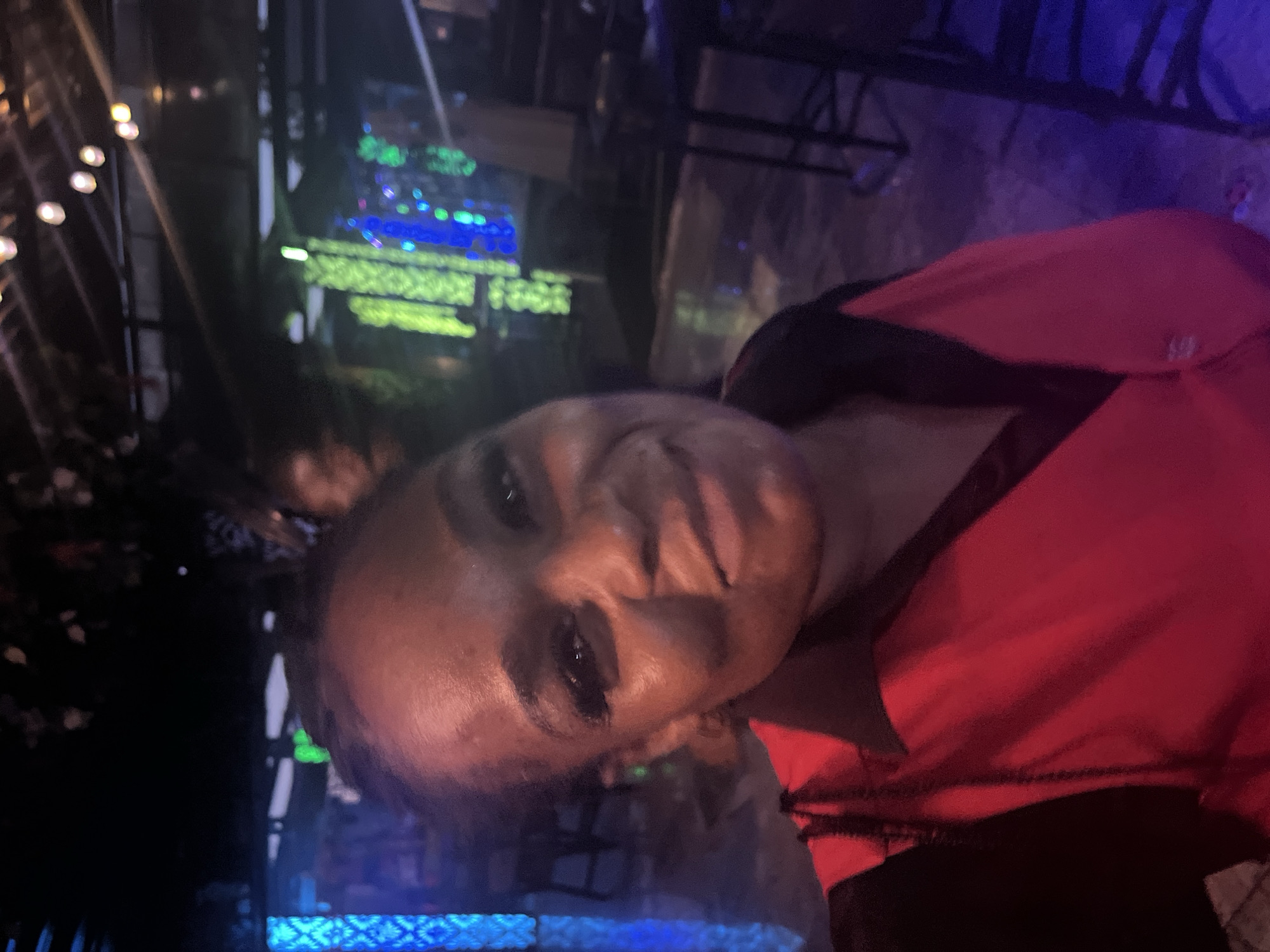 A single individual, an adult female with a dark complexion, is prominently featured in a close-up, chest-up view. She is depicted with a subtle smile, directly engaging the viewer. Her hair is pulled back, and she is wearing a red garment with a black collar and black shoulder/sleeve sections, suggesting a uniform or casual top. The background indicates a dimly lit indoor or covered outdoor commercial establishment, characteristic of a bar, lounge, or nightclub. Ambient lighting is provided by various artificial sources: a tall, vertical structure on the far left displays a blue, patterned illumination. On the right, multiple vertical columns emit bright green light, interspersed with some blue lights further back. Overhead, soft, warm-toned circular lights are visible against a dark ceiling structure. Minimal furniture is discernible, including a dark tall table with a stool on the right side of the frame, and a partial view of another table and chair on the far left. The floor appears dark and slightly reflective in areas. The overall impression is one of a vibrant, evening leisure environment.