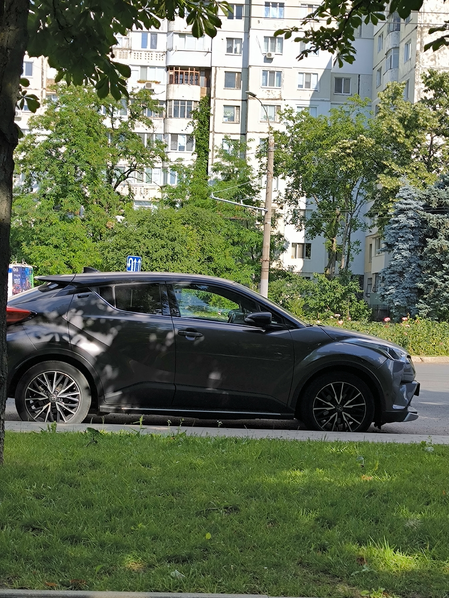 A dark gray crossover vehicle is parked on the side of a street, partially obscured by tree branches and foliage. The car has a sporty design with black and silver alloy wheels. Behind the car, a tall, multi-story apartment building with a light-colored facade is visible. Green trees and bushes surround the building and line the street. A blue traffic sign with white arrows is mounted on a pole above the car. The foreground consists of a strip of green grass. The scene appears to be a typical urban residential area in Chișinău, Moldova, on a sunny day.