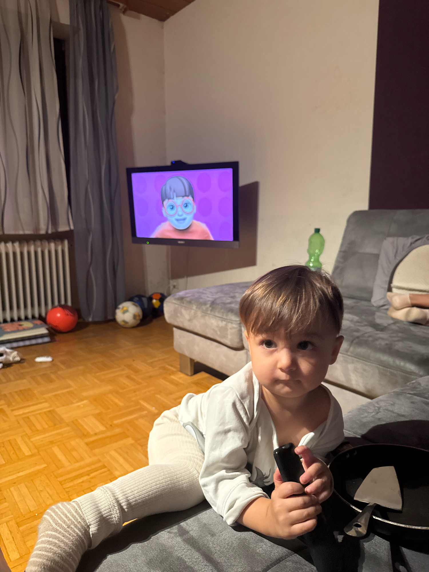 In an indoor setting, a young child with light brown hair and brown eyes is seated on a gray sofa, holding a black remote control. The child is wearing a white long-sleeved shirt and light-colored leggings with striped socks. A black frying pan with a spatula rests on the sofa next to the child. In the background, a television screen displays a cartoon character with blue skin and pink glasses against a purple backdrop. To the left of the television, gray curtains hang over a radiator. The room features a wooden floor, and several soccer balls are scattered on the floor near the radiator. A bright green plastic bottle is visible on the sofa behind the child. The overall lighting suggests it is evening. This scene is consistent with a domestic interior in Überherrn, Germany.