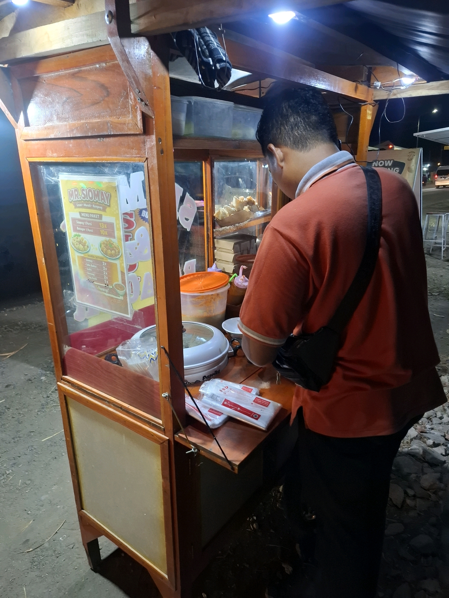 The image shows a person in an orange shirt standing in front of a wooden food cart at night. The food cart has a glass display case showing some food items and a menu board that reads "MR. SIOMAY". The person appears to be interacting with the cart. The environment is dimly lit, suggesting it is an outdoor street setting. The ground is covered with gravel and dirt. There are some rocks and debris on the ground to the right. In the background, there are some lights from buildings and vehicles, indicating a city environment. There is a visible sign that says "NOW" suggesting it is an advertisement.