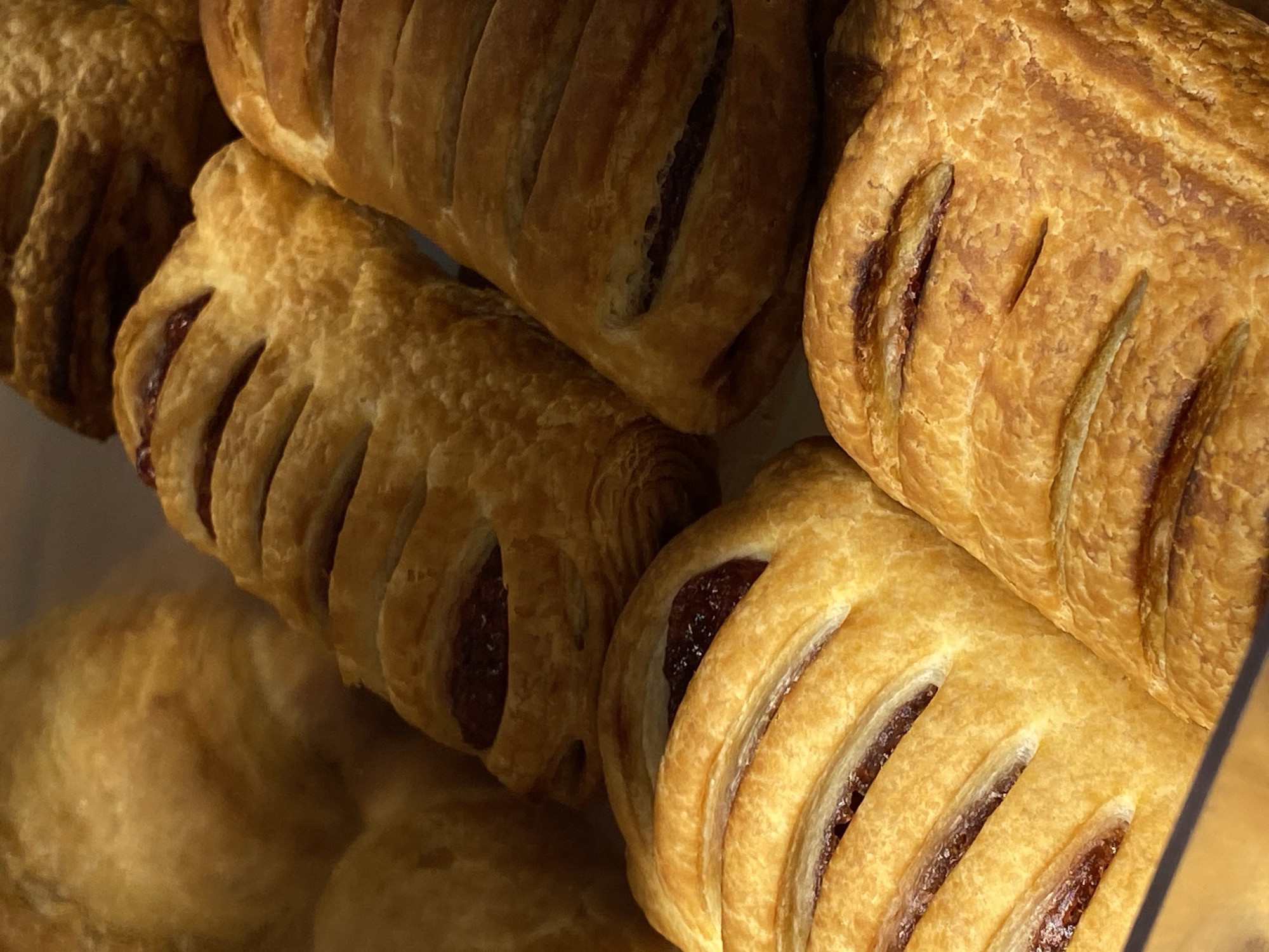 A close-up shot depicts several pastries. The pastries have a golden-brown, flaky crust and are filled with a dark-colored jam or fruit filling visible through cuts in the pastry. The pastries are arranged in a pile, with some partially overlapping others. The pastries are indoors, perhaps in a bakery or a shop, likely in Votkinsk, Russia. The scene includes no people.