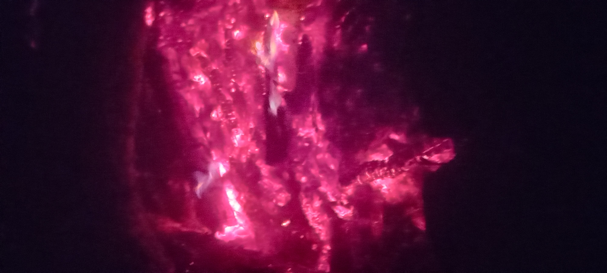 The image presents a detailed view of highly incandescent material, predominantly in shades of fuchsia, magenta, and crimson, set against a deep black backdrop. The scene is situated in the city of Kryvyi Rih, Ukraine. Brightest luminosity is concentrated in the upper-central and mid-left areas, where vivid white and light pink hot spots are visible amidst the darker red fragments. The texture of the glowing elements suggests a turbulent, granular, or crystalline structure, indicative of intense heat, possibly from burning embers or molten substances. Towards the lower-left, an elongated, darker object, consistent with a charred piece of wood or debris, is visible, partially obscured by and reflecting light from the surrounding glowing matter. No human subjects, animals, or specific architectural features are present within the frame.