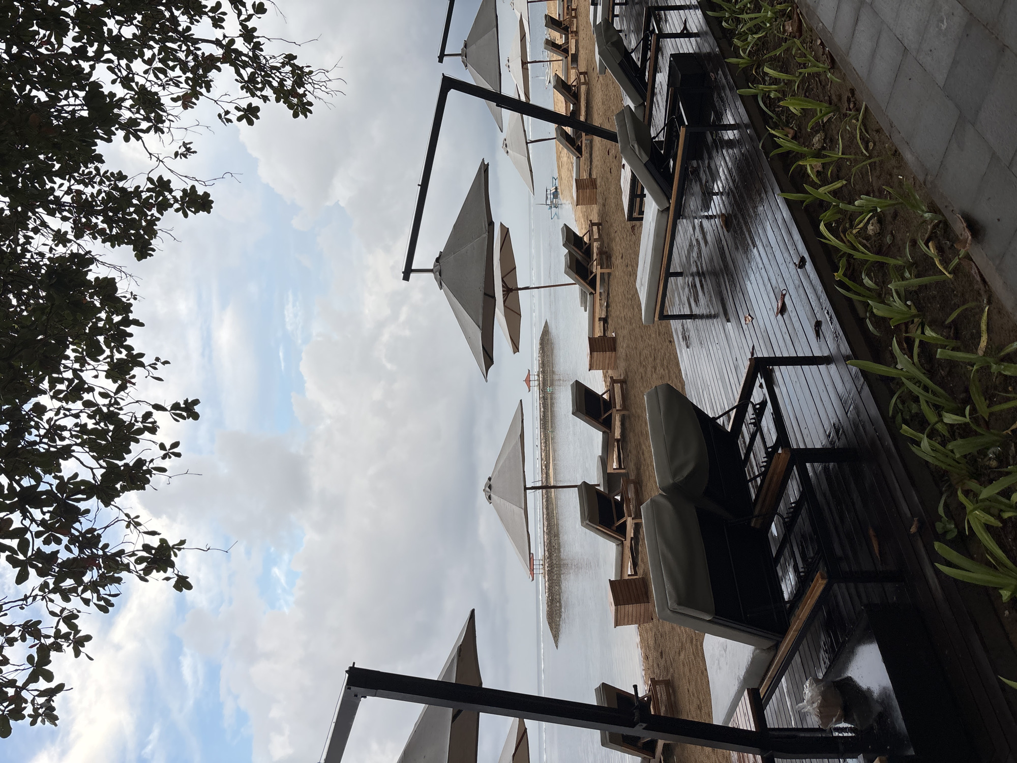 The image depicts a deserted beach in Denpasar, Indonesia, under a cloudy sky. In the foreground, several lounge chairs with gray cushions are arranged on a wet wooden deck. To the left, a black metal umbrella frame extends upwards, supporting a large gray umbrella. Beyond the deck, the sandy beach is dotted with more lounge chairs and gray umbrellas. The calm sea stretches to the horizon, with a breakwater and small shelters visible in the distance. A few boats are anchored in the water. The overall atmosphere is tranquil and somber, suggesting a recent rain shower.