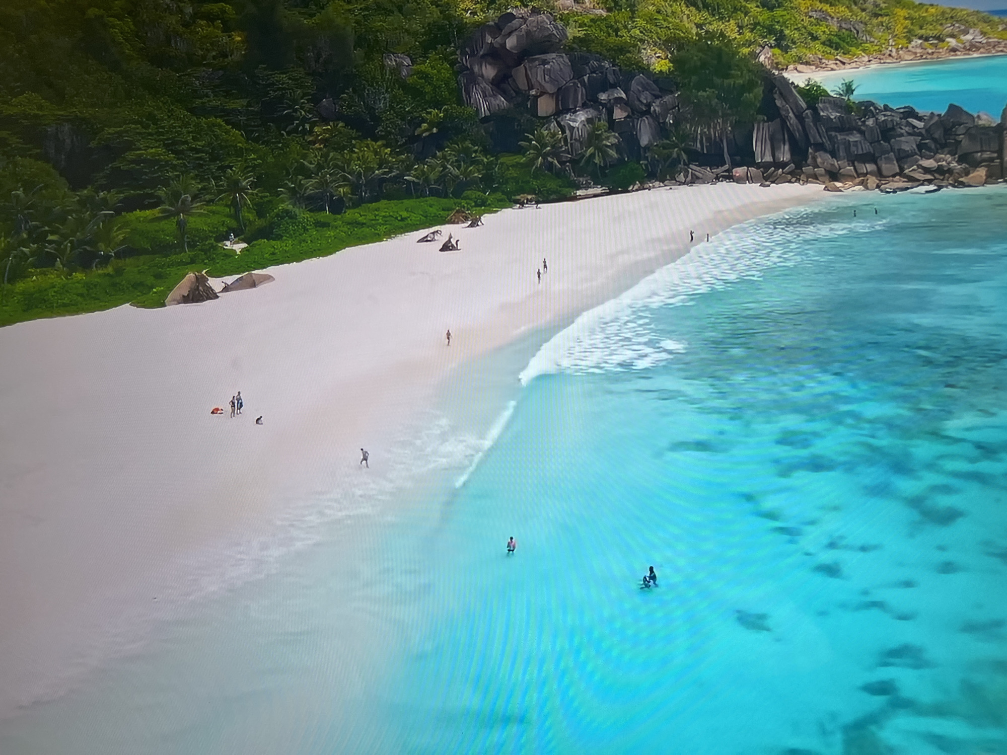 The image shows a tropical beach with white sand and turquoise water. A rocky outcrop borders the beach, covered in green vegetation. Several people are on the beach and in the water, some swimming and others walking along the shoreline. The location is not in Basingstoke and Deane, United Kingdom; it is a tropical beach.