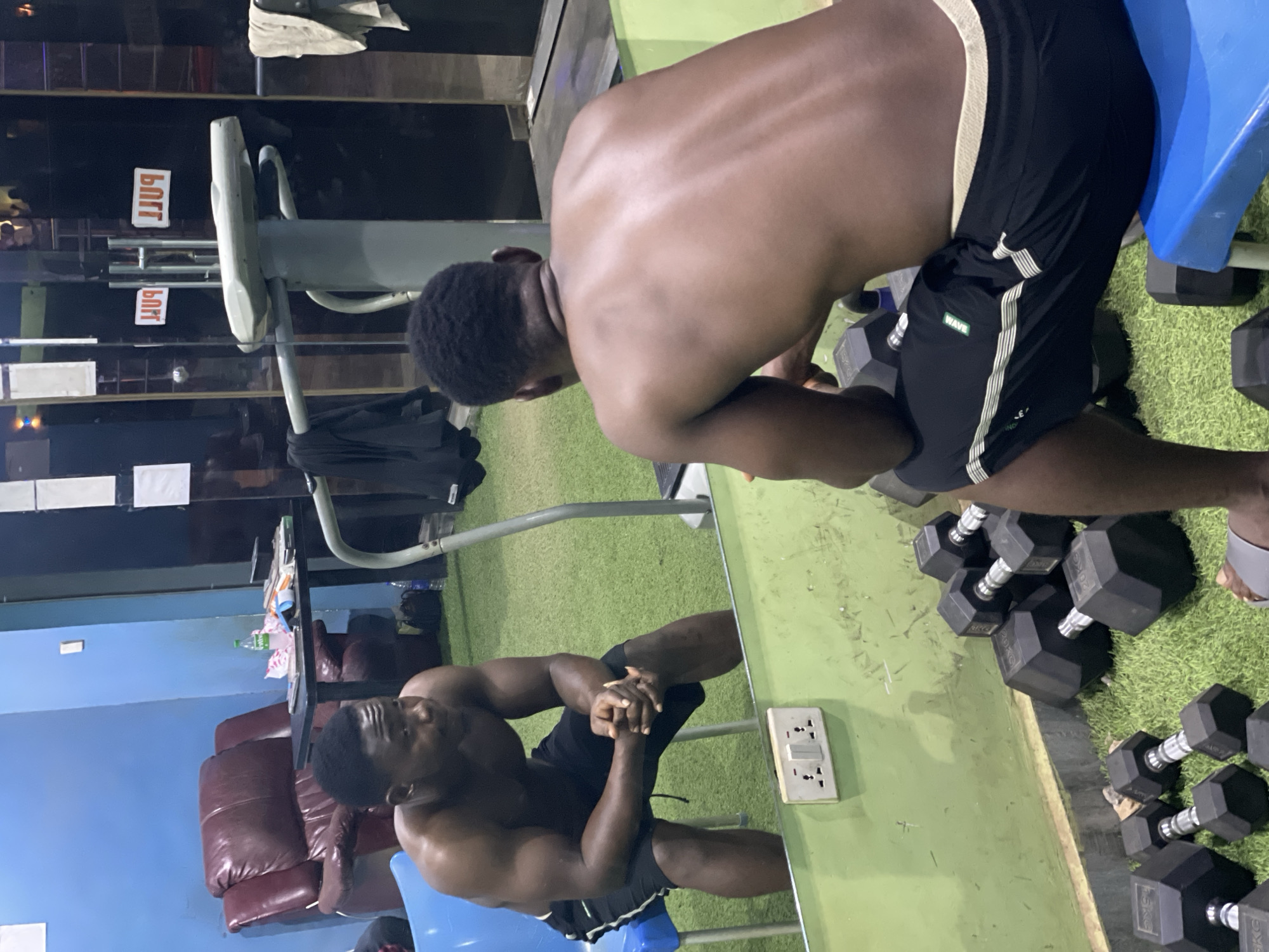 A shirtless muscular man with dark hair is seen in a gym. He is wearing black shorts with white stripes and is seated on a blue chair. The room contains multiple black hexagonal dumbbells on the floor. A reflection of the man can be seen in a shiny surface. The gym includes a treadmill and upholstered chairs. The location is Akwanga, Nigeria.