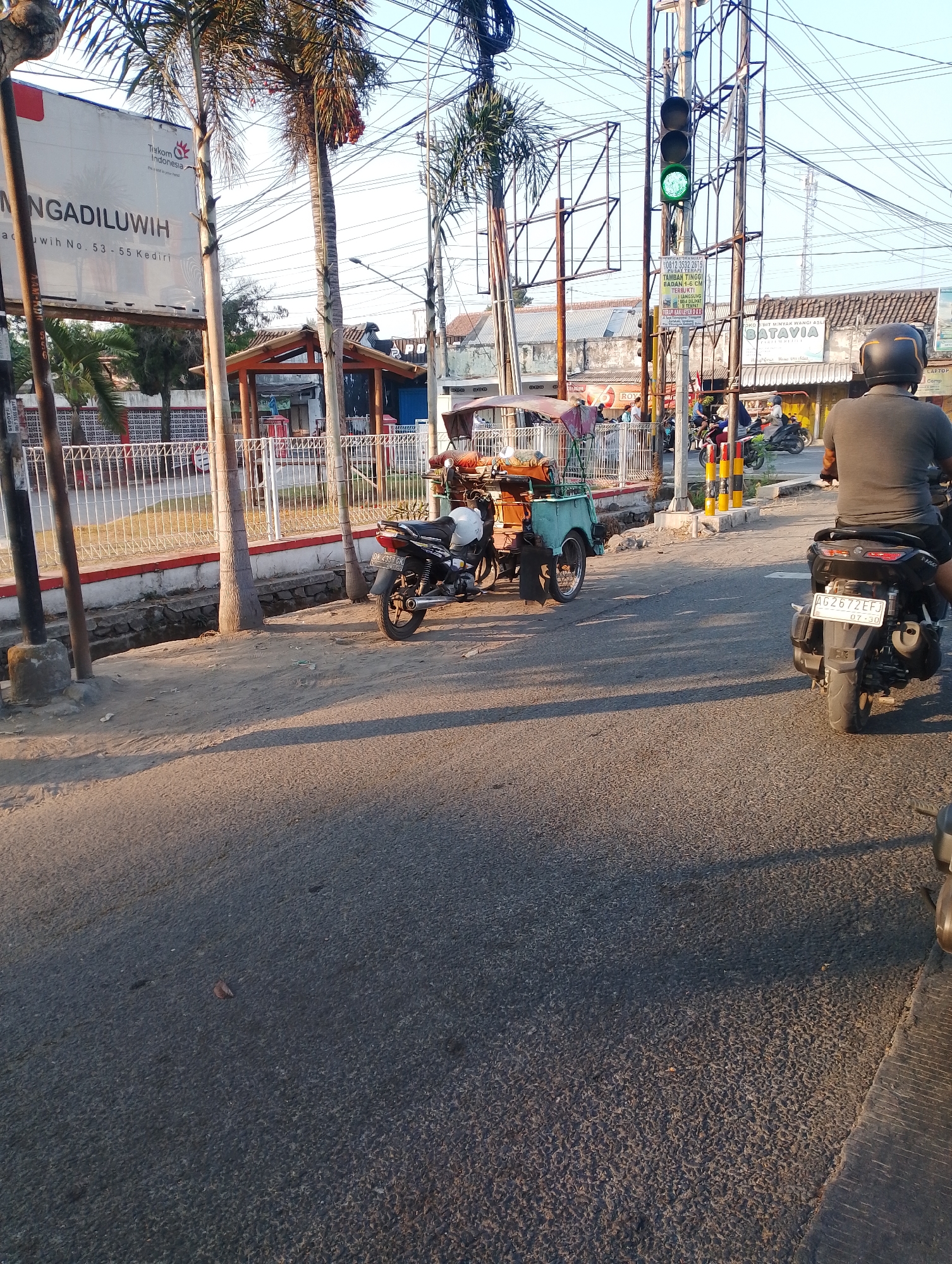 The image depicts a street scene in Dukuh, Indonesia. A traffic light is visible, showing a green signal. Several palm trees line the sidewalk, which is adjacent to a road. On the road, a three-wheeled vehicle, possibly a becak, is parked. In the foreground, a person on a motorcycle is seen from behind, with another motorcycle and a becak further down the street. Buildings with signs are visible in the background. The lighting suggests it is either early morning or late afternoon.