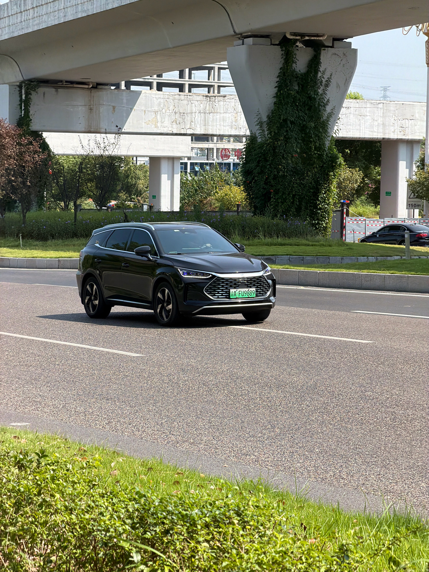 A black BYD Tang SUV with a green license plate reading "A-FU9889" drives on an asphalt road. The car is positioned slightly to the left of the frame's center, heading towards the right. Behind the SUV, a second black car is visible further down the road. The scene is set under a large concrete overpass, supported by sturdy pillars. Green ivy climbs up one of the pillars. The environment includes manicured green grass and bushes along the roadside, with trees in the background. A sign indicating "150m" is visible to the right. The sky is clear and blue. The location is identified as Jiangjin District, Chongqing City, China, likely near a transportation hub or elevated roadway.