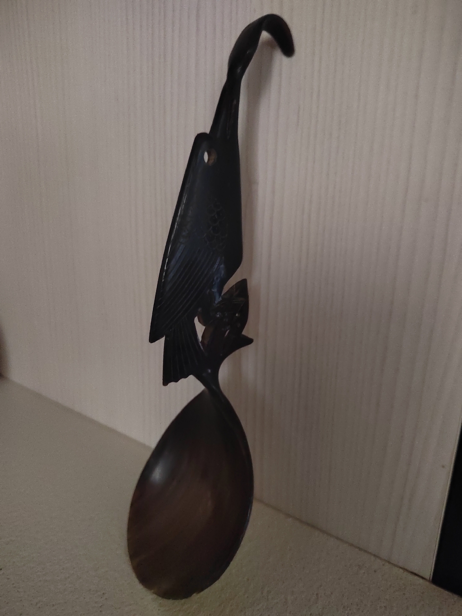 A carved wooden spoon with a bird as the handle rests on a beige surface against a beige wall with vertical lines. The spoon is dark brown and the bird appears to be an eagle or hawk with detailed feathers. The bird is perched on what seems to be a branch which then transitions into the bowl of the spoon. The image is taken from a slightly low angle. The spoon is the primary object in the scene and is positioned vertically.