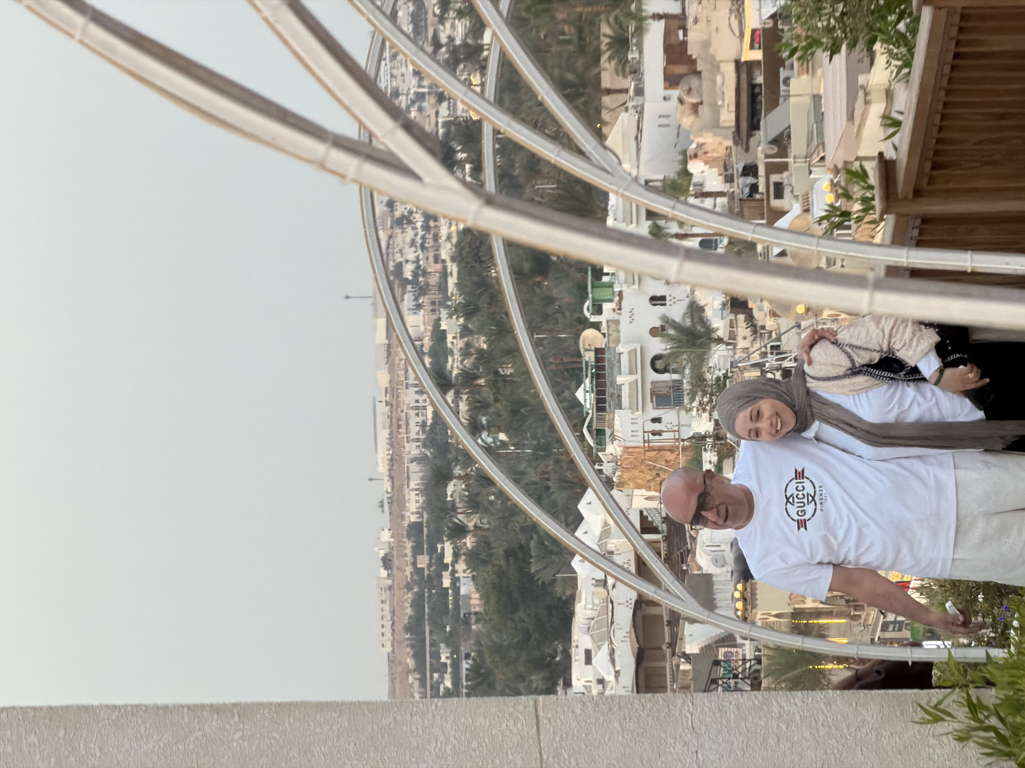 A man and a woman are posing for a photo in Sharm El Sheikh, Egypt. The man is bald, wearing sunglasses, a white t-shirt with a Gucci logo, and white shorts. The woman is wearing a grey hijab and a light-colored jacket. They are standing on a wooden balcony with a decorative metal archway in the foreground. In the background, there are white buildings with flat roofs, palm trees, and distant hills. The sky is overcast.