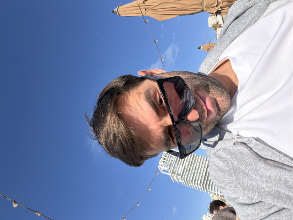 The image captures a male individual, appearing to be in his 30s-40s with short brown hair and light facial stubble, taking a selfie-style photograph. He is wearing large black-framed sunglasses, a white t-shirt, and a grey zippered hoodie, and looks towards the camera with a slight downward gaze and subtle smirk. The perspective is from a low angle, with a clear, bright blue sky dominating the upper portion of the frame.
The outdoor setting is identifiable as Tel-Aviv, Israel. In the mid-ground to the left of the man, a distinctive white multi-story building with terraced architecture, consistent with The Norman Tel Aviv hotel, is visible. String lights with exposed bulbs are strung across the upper left background, suggesting an outdoor patio or dining area. To the right of the man, a large, closed brown canvas sun umbrella is prominent, with another partially visible behind it, along with white building facades. The scene is brightly lit by natural sunlight. A portion of another person's head with dark hair is visible in the far left background.
