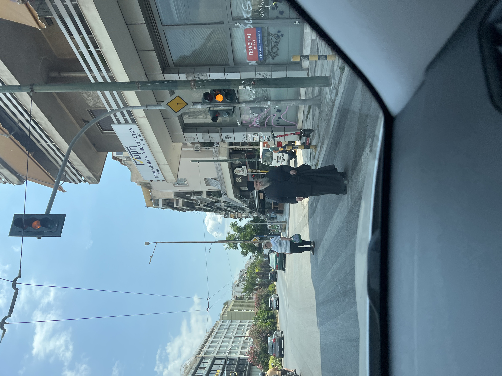 In Kallithea, Greece, on a sunny day, a street scene unfolds with pedestrians and traffic. A man in traditional black clerical attire with a long beard is walking across a crosswalk, his gaze directed forward. To his left, a woman in a striped shirt and dark pants also crosses the street, carrying a green bag. A traffic light above them shows an orange signal. Behind them, a white van and a red electric scooter are parked near a building with "FOR SALE" signage. The street is paved, and buildings line the opposite side. The perspective suggests the image is taken from inside a vehicle.