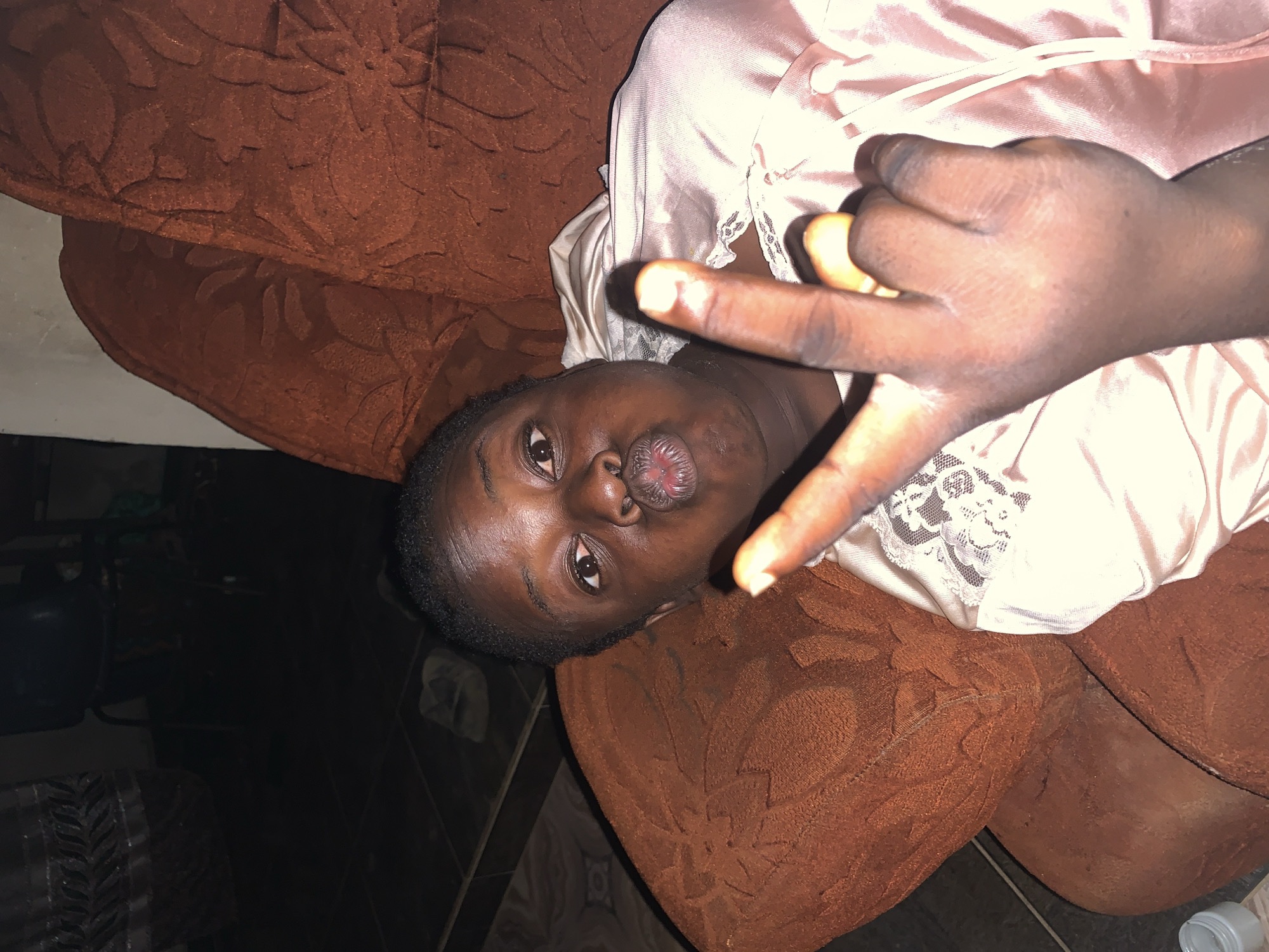 A dark-skinned person is lying on a brown patterned sofa or couch. The person is wearing a pink satin top with lace detailing around the collar. The person's face is turned towards the camera and their lips are pursed. The person is holding up a hand with the index and middle fingers extended in a "peace" sign or "victory" gesture. The background is dark, with a tiled floor and the suggestion of other furniture or objects partially visible.
It's impossible to verify the location as Agban, Nigeria, based on this picture.