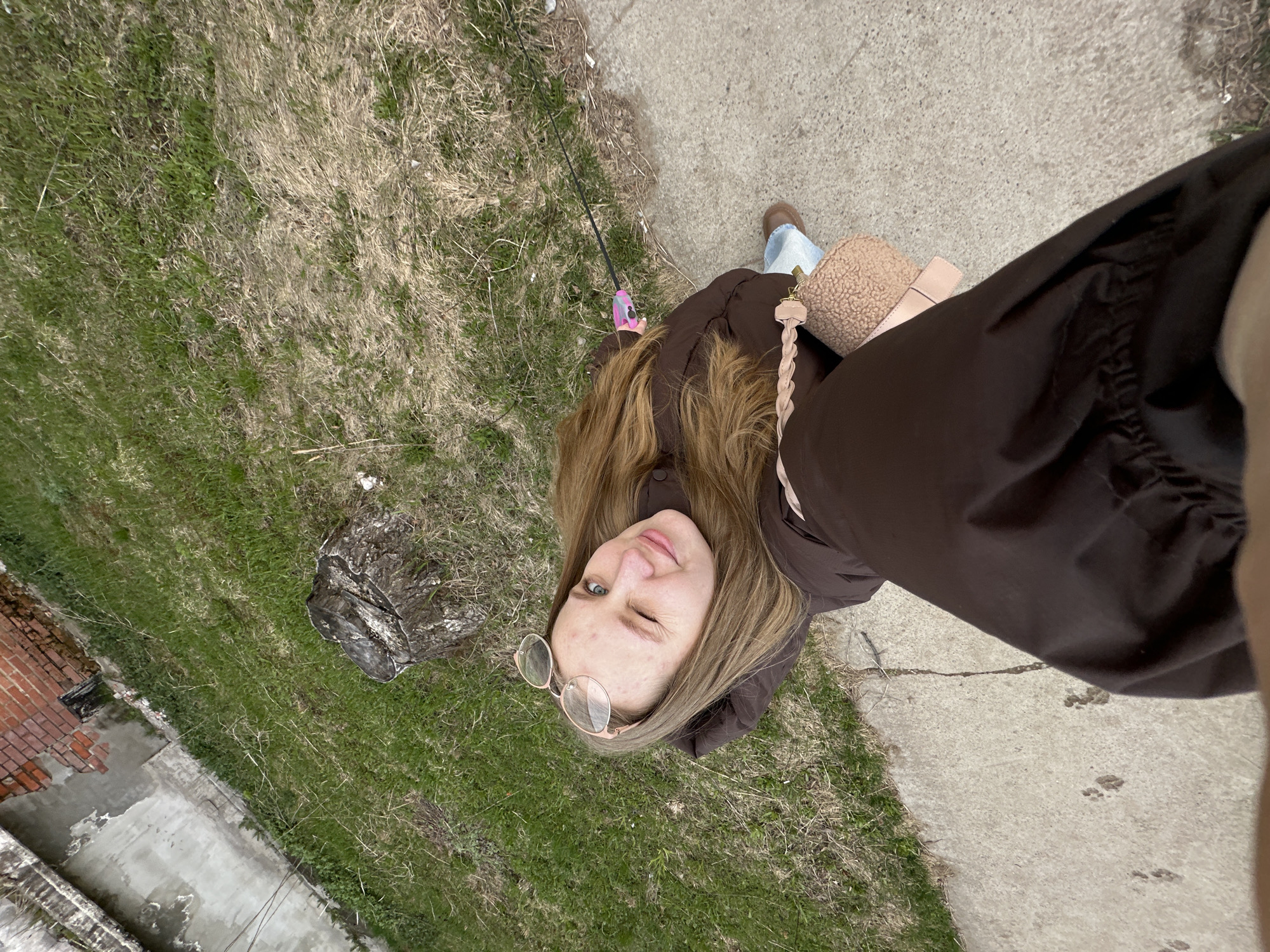 The image features a young woman outdoors in Chaikovsky, Russia, captured in a playful selfie or close-up shot. Her head is tilted, and she is winking at the camera, conveying a friendly and casual mood. She has long, light brown hair and wears clear, round-framed glasses. Her attire includes a dark brown puffer jacket, light-wash jeans, and brown shoes or boots. A light-colored, possibly teddy-textured, shoulder bag with a braided strap hangs over her shoulder.
To her right, a concrete sidewalk shows several small animal paw prints, suggesting she is accompanied by a pet. A thin black leash extends from her left, connected to a small pink object, likely a dog waste bag dispenser or a toy, further indicating a dog walk. The ground to her left is a mix of patchy green and dry brown grass, with a dark, weathered tree stump or log nearby. In the lower-left background, there is exposed brickwork and another section of concrete or paved ground, suggesting an urban or suburban setting. The scene is illuminated by natural daylight, indicating a bright or overcast day without harsh shadows. No visible text is present in the image.