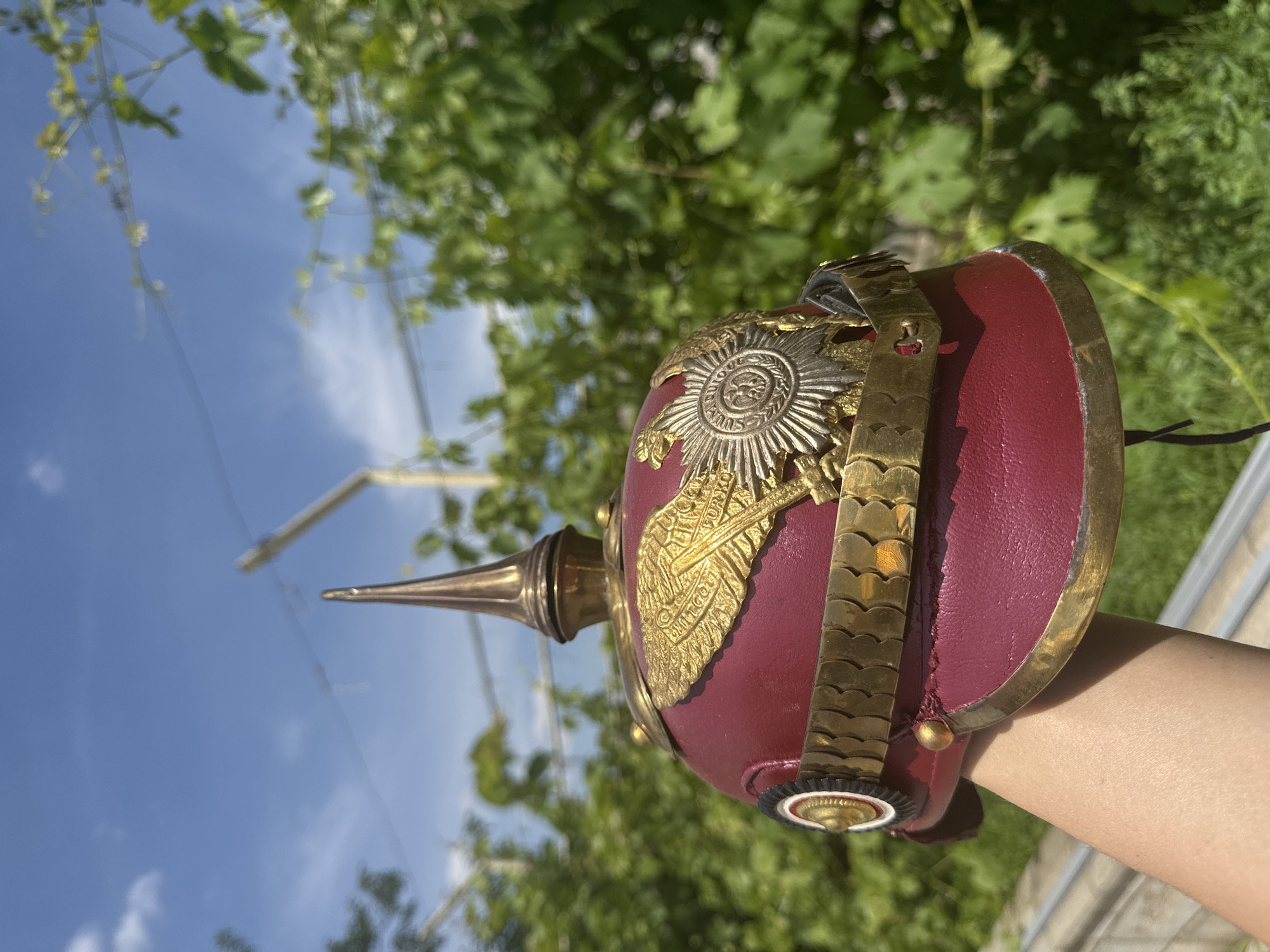 A close-up, sideways shot shows a red, ornate helmet held by a person's arm, with a bright blue sky and green foliage in the background. The helmet, likely a historical military or ceremonial piece, is adorned with metallic detailing. A prominent feature is a silver emblem with radiating points, possibly a sunburst or star, centered on the helmet's front. Below this, a golden, wing-like ornament is attached, possibly symbolizing an eagle or a feather. A segmented gold band encircles the helmet. A pointed metallic spike extends from the top. The helmet's brim has a brass or gold-colored trim. The person's arm, lightly tanned, is visible supporting the helmet. The setting appears to be outdoors during the day, with clear blue sky visible with some wispy clouds. The abundant green foliage suggests a natural environment, possibly a garden or park. The lighting indicates it's daytime, likely sunny. The image evokes a sense of historical interest or admiration for the artifact. No visible text is discernible.