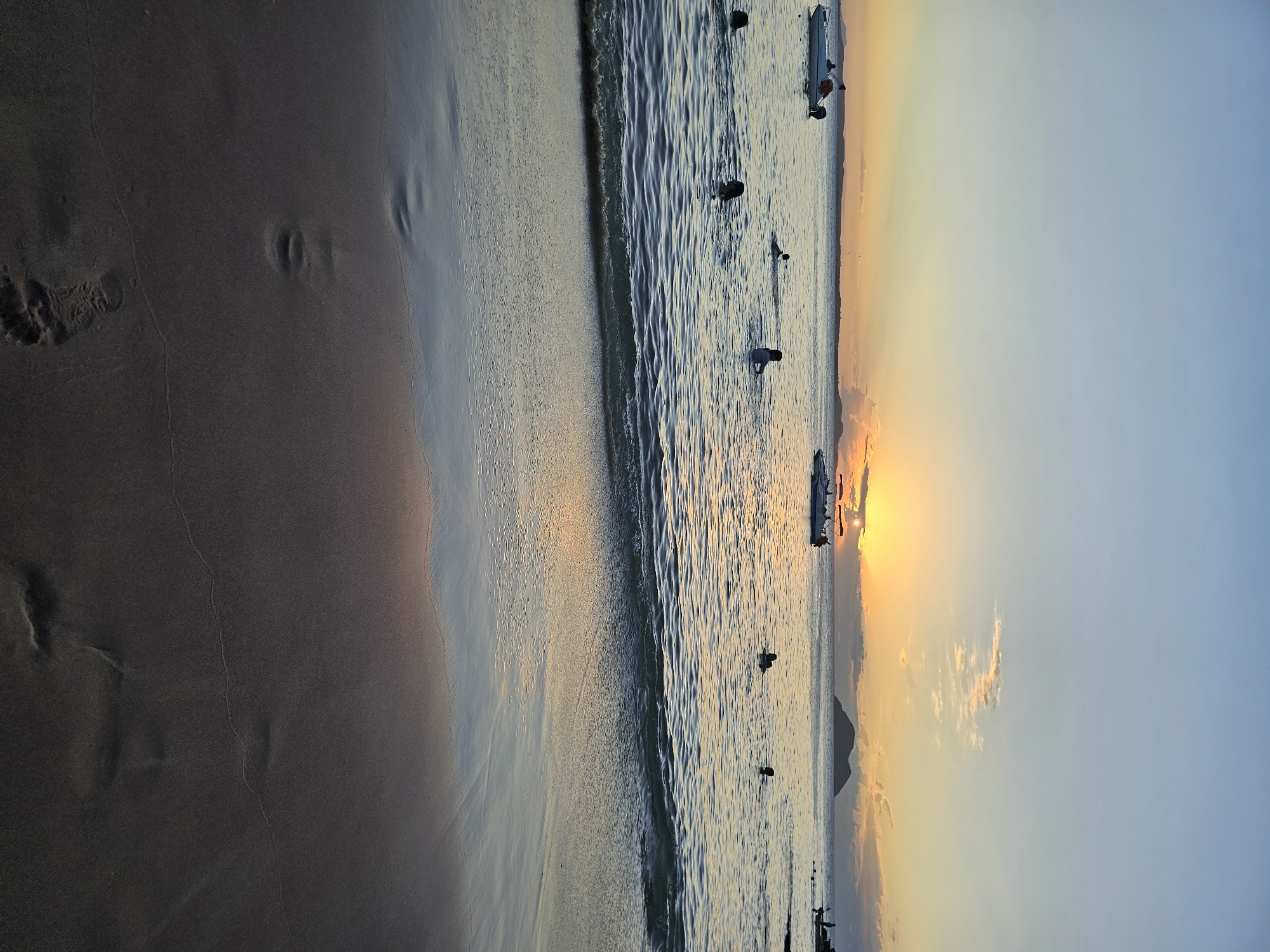 Here's a forensic description of the image:
**Scene:** A beach at sunset in Armação Dos Búzios, Brazil.
**Environment:** The sky transitions from a light blue to orange-yellow at the horizon. The sun is setting over the water. The beach is made of dark sand, with visible footprints near the shoreline.
**Objects:** Small boats are present on the water.
**People:** Several people are in the water.
**Actions:** People are swimming in the water and presumably enjoying the sunset. The waves are washing onto the sand.