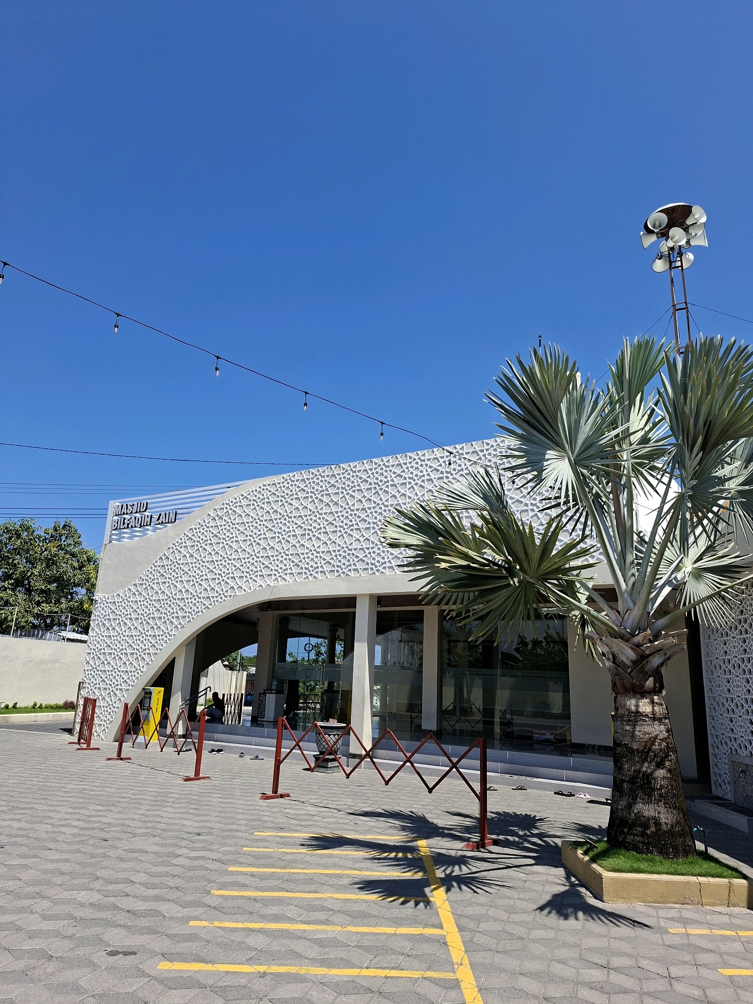 The image shows the exterior of a modern mosque with a curved, white facade adorned with a geometric pattern. A prominent palm tree stands in front of the building. The mosque has large glass windows and doors, and text above the entrance reads "MASJID BIL FAQIH ZAIN." The ground in front of the mosque is paved with grey interlocking bricks, marked with yellow lines that appear to indicate parking or pedestrian pathways. Several red barrier stands are set up on the paved area. In the background, a clear blue sky is visible, with some power lines and a speaker system mounted on a pole. A few pairs of flip-flops are visible near the entrance. The location is identified as the city of Branta Tinggi, Indonesia.