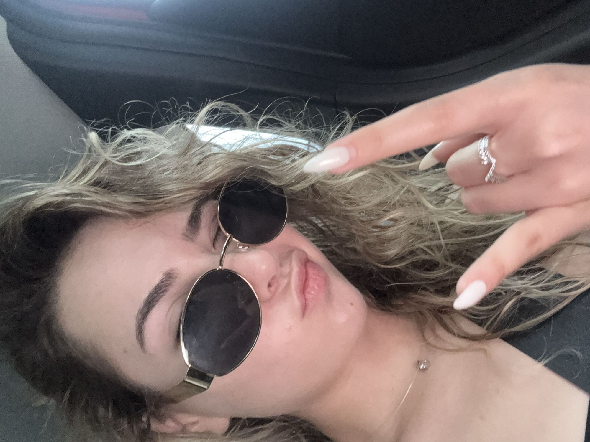A close-up portrait shows an individual, likely female, with fair skin and wavy, light brown hair, wearing dark, oval-shaped sunglasses with gold frames. The eyebrows are dark and arched, and the lips are pursed in a pout. The subject's left hand is raised, performing a "rock on" gesture with the index and pinky fingers extended. The nails are long, almond-shaped, and coated with a light, translucent polish. A silver-toned ring with a wavy, textured design is worn on the left ring finger. A delicate, thin necklace with a small, spherical pendant is visible on the neck. The individual is wearing dark attire, evidenced by a black strap across the collarbone area. The background is indistinct and dark, consistent with the interior of a vehicle. No visual cues are present to determine a specific location within the city of Sofia, Bulgaria, or any other geographic area.