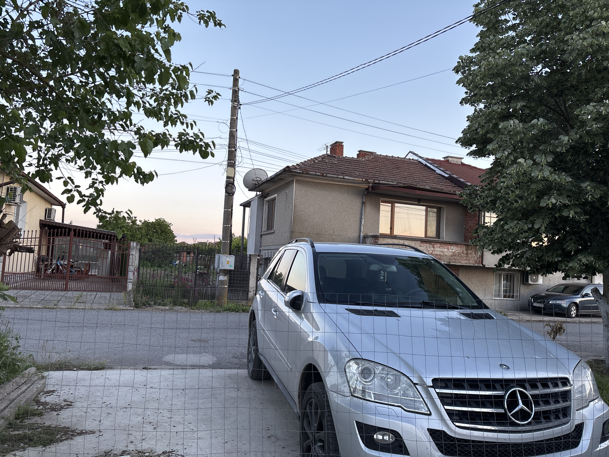 The image captures a street scene in an urban location. A silver Mercedes-Benz SUV is parked in a driveway behind a wire fence. In the background, a two-story house with a tiled roof stands next to another building partially obscured by foliage. Electrical wires and a utility pole are visible against a pale blue sky. The street is paved and there are fences along the sides of the properties. A second dark-colored car is parked in front of the house to the right of the frame. Vegetation is present on both sides of the frame, with a large tree on the right and branches visible on the left.