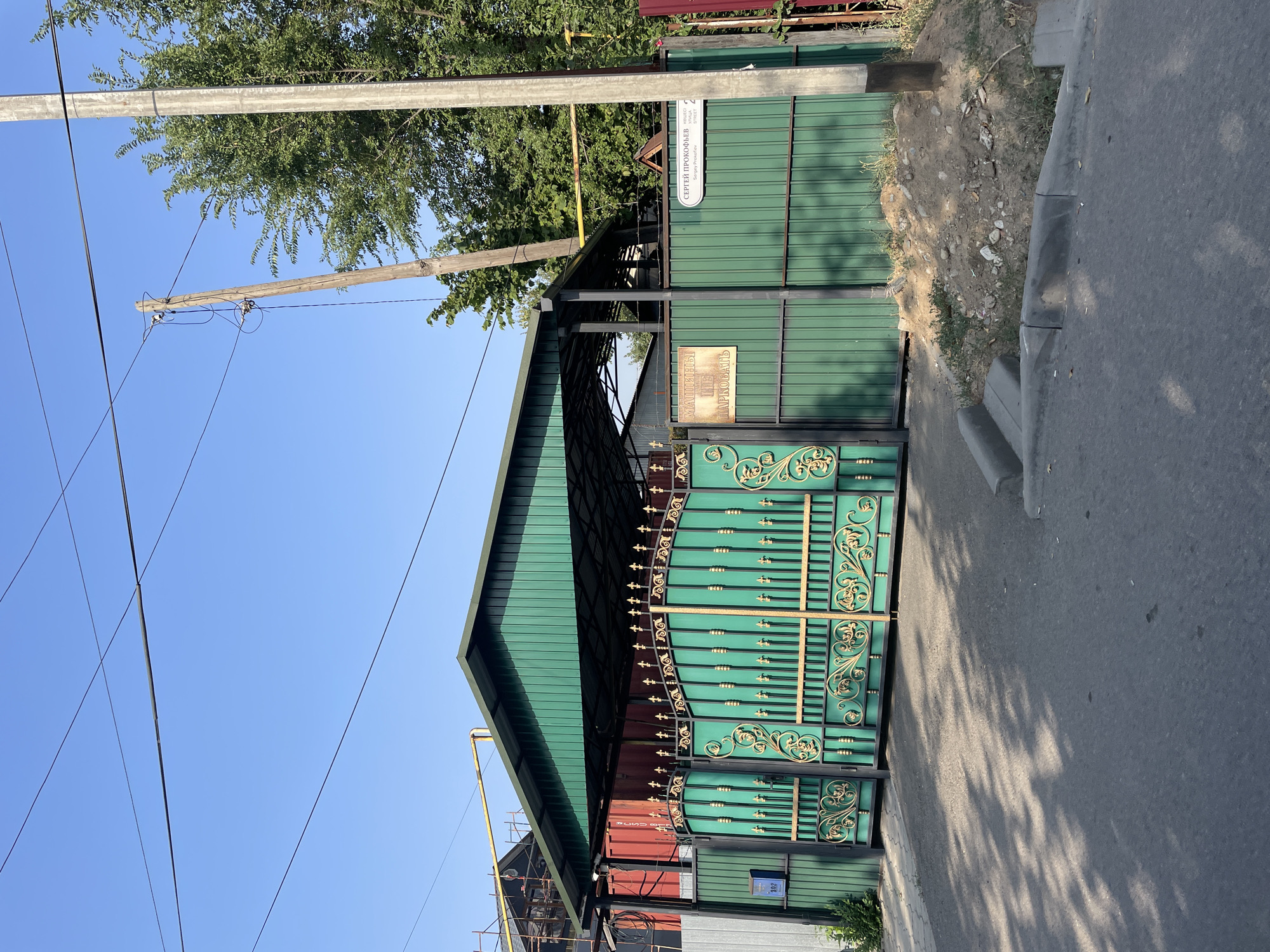 A green metal gate with ornate gold detailing is the central focus of the image, situated in front of a green metal fence. Above the gate, a green metal roof supported by dark metal beams provides cover. To the left of the gate, a blue mailbox with the number "302" is visible on the green fence. A sign with text in Cyrillic is affixed to the gate, and another sign with Cyrillic text is attached to the fence to the right of the gate. Behind the fence, industrial-looking structures with red shipping containers and scaffolding are partially visible. Trees and power lines dominate the background, with a wooden utility pole and a concrete pole standing prominently. The scene appears to be on a street in Almaty, Kazakhstan, indicated by the Cyrillic signage. The road in the foreground is asphalt, with a concrete curb and sidewalk. The sky is clear and blue.