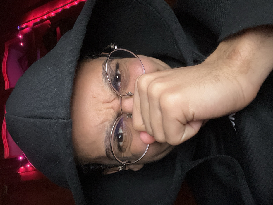 A person wearing a black hoodie and glasses is shown with a clenched fist near their face. The individual's eyes are visible between the glasses and the fist. Red string lights are present in the background. The image is slightly dark and appears to be taken from an unconventional angle.