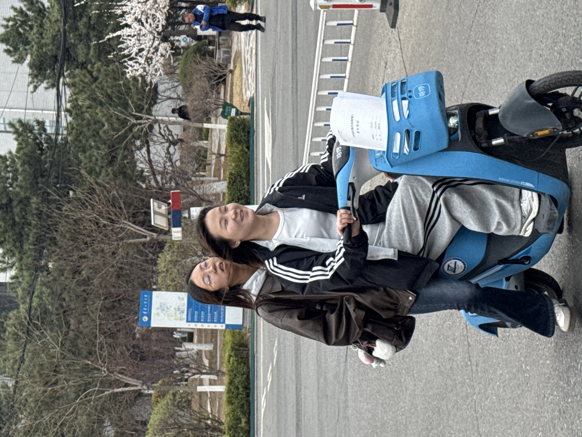 Two young women are riding a blue scooter on a paved road in Zhongtun, China. One woman is wearing a brown jacket, jeans, and glasses. The other woman is wearing a black jacket with white stripes, a white shirt, and gray pants. They are both smiling. The scooter has a blue basket on the front. There are trees and bushes in the background, along with a sign and a building. Another person is seen in the distance in the upper left corner.