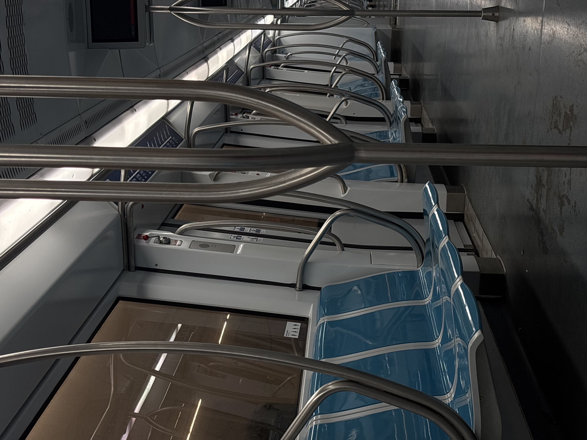 The interior of a subway car is captured in this tilted, high-angle shot. Rows of blue, contoured seats line the car, separated by metal armrests. Overhead, metal handrails run the length of the car. Windows are visible, but the view outside is obscured. The floor is a dark, solid color. There are no people visible in the scene. The car appears to be clean and modern. This is most likely the interior of a train on the Rome Metro, in Rome, Italy.