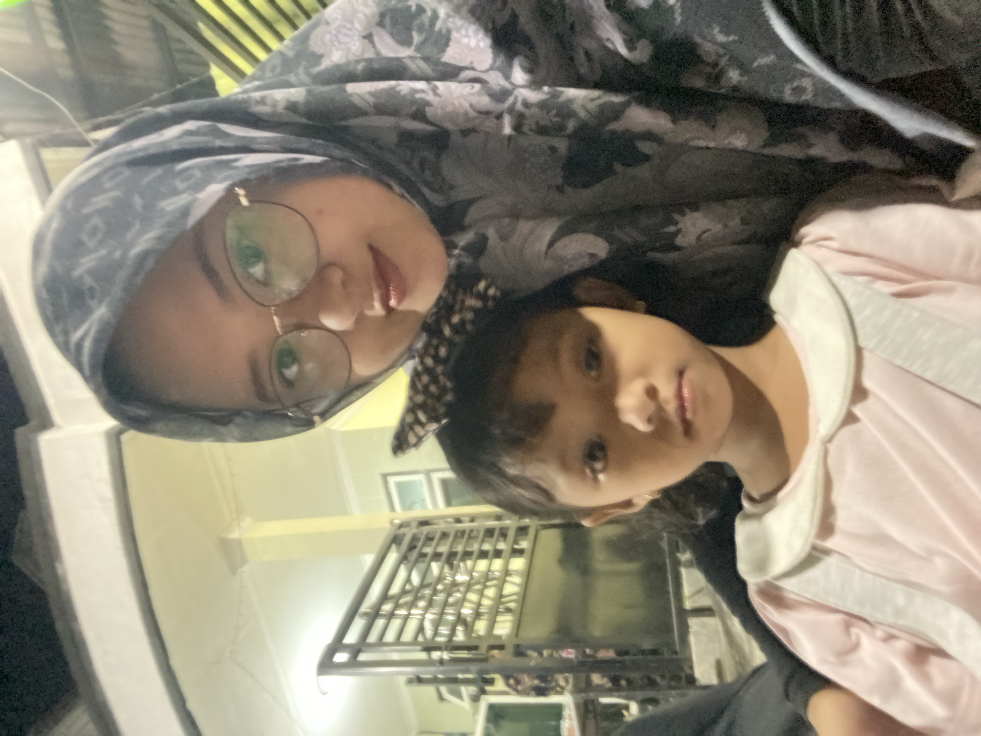 The image shows two people, likely a woman and a child, indoors. The woman is wearing a headscarf and glasses. The child is wearing a light pink garment with a white collar and a spotted headband. The background features a metal structure, possibly part of a bed frame or furniture, along with indoor lighting and glimpses of what appears to be a room or building interior. Based on the metadata, the location is Gelam, Indonesia.