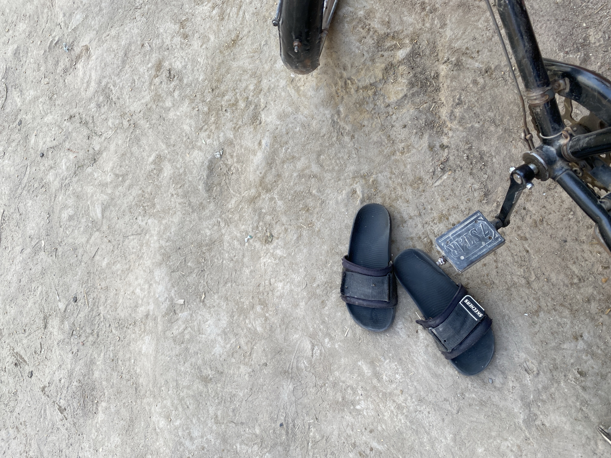 The image presents an overhead view of a pair of black slide sandals and parts of a bicycle resting on a dusty, light-colored ground surface in Chak 166 GB, Pakistan.
In the center-right, a pair of black slide sandals is placed on the ground. The strap of the right sandal clearly displays the brand name "SKECHERS" in white lettering, oriented upside down. To the right and upper right of the sandals, components of a bicycle are visible. These include a section of a black front wheel or mudguard at the top edge, and parts of the black bicycle frame, chainring, and a pedal towards the right. The bicycle pedal has the text "7 STAR" embossed on its surface.
The ground is a mix of light gray and beige dirt, appearing dry and somewhat uneven, with small pieces of debris and faint markings scattered across its surface. The bright, even lighting suggests it is daytime. No people are present in the frame, but the arrangement of the sandals and bicycle implies recent human activity, such as someone having dismounted the bike or preparing to ride, and leaving their footwear on the ground. The scene captures a simple, everyday outdoor moment in a dusty environment.