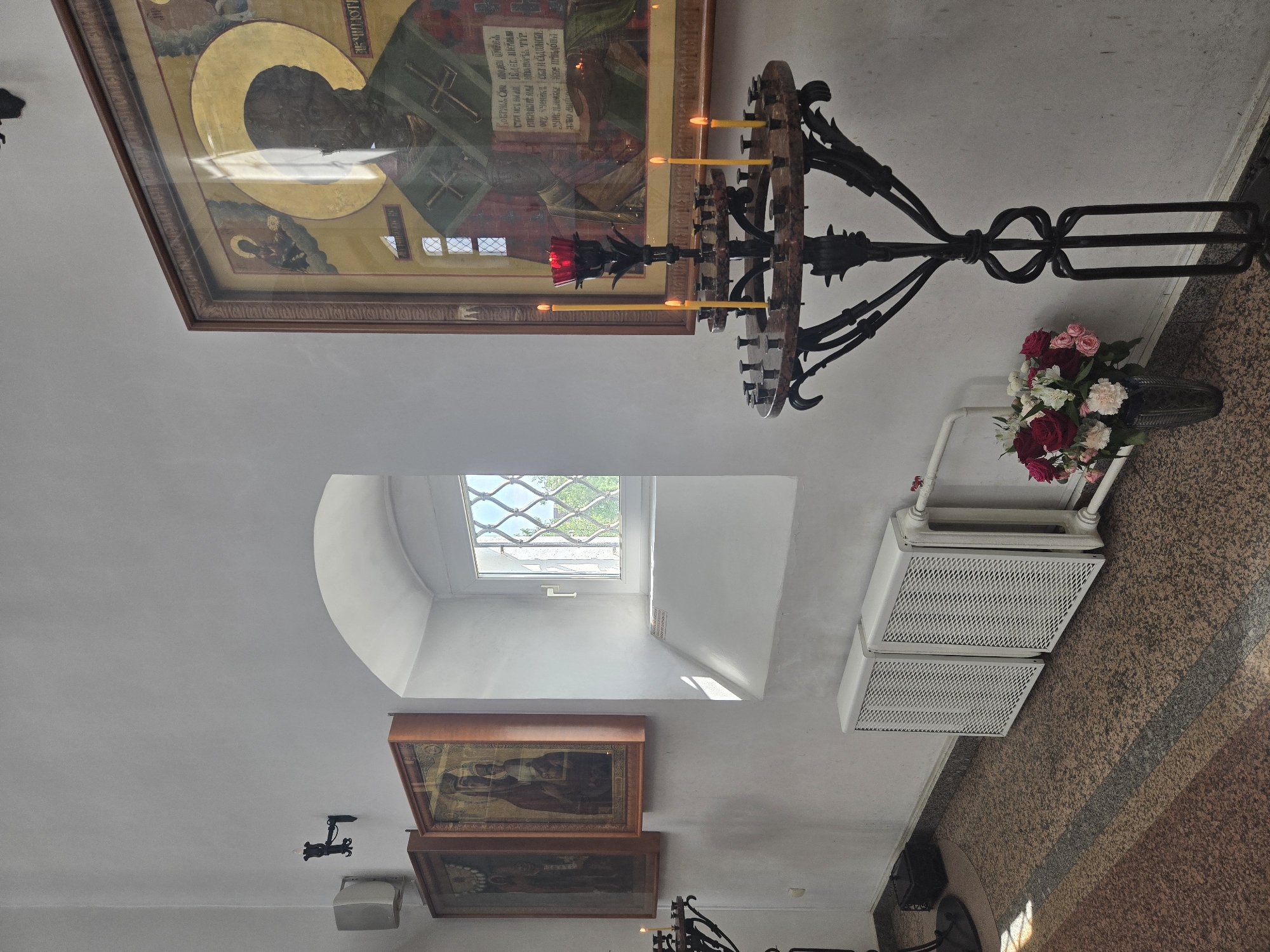 The image shows the interior of a building, possibly a church, in Dolgoprudny, Russia. On the upper portion of the image, a large framed icon is visible, along with an ornate, circular chandelier holding lit candles. The walls are white. A rounded window provides natural light. Below the window, smaller framed images are displayed. A white radiator and a vase of flowers rest on the floor. The floor is covered in patterned tile.