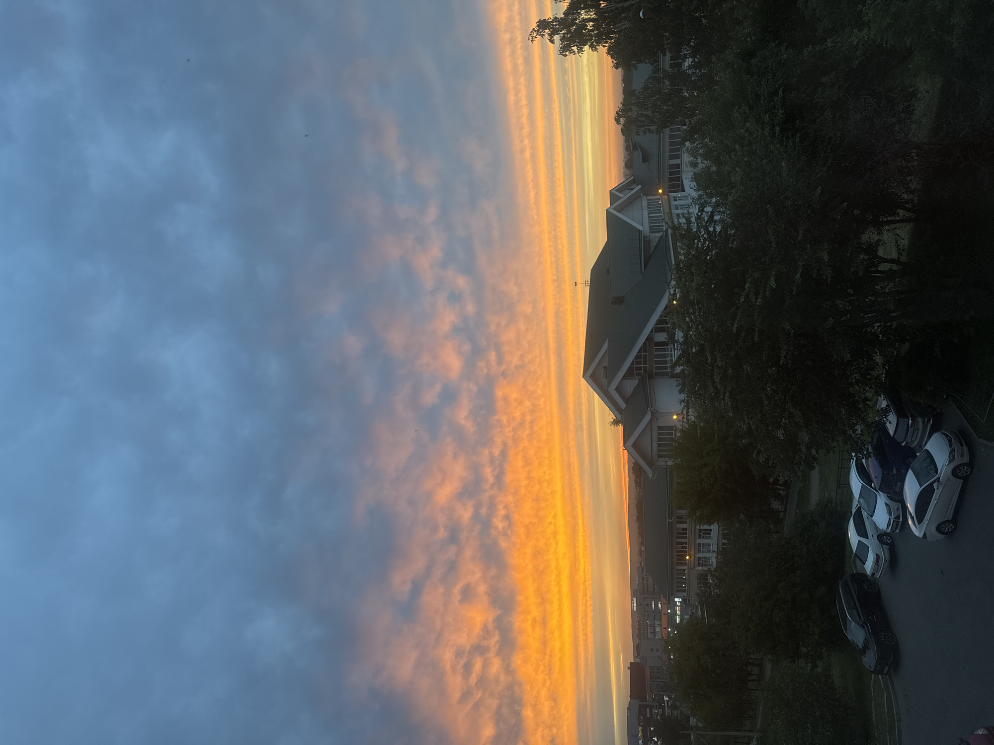 The image captures a twilight scene in the city of Irkutsk, Russia, likely during late summer or early autumn. A dramatic sky dominates the upper portion of the frame, with vibrant streaks of orange and yellow clouds illuminated by the setting sun, contrasted against a cooler, blue-grey expanse above. Below the sky, a suburban or residential area is visible. Several multi-story buildings with pitched green roofs are prominent, appearing to be apartment complexes or institutional buildings. Some of these buildings have visible windows and balconies. In the foreground, a parking area is occupied by several cars, including a dark SUV, white sedans, and a dark-colored sedan. Trees and other greenery surround the buildings and the parking lot, some of which are casting shadows due to the low light conditions. The overall atmosphere suggests a peaceful evening in a developed urban or suburban setting.