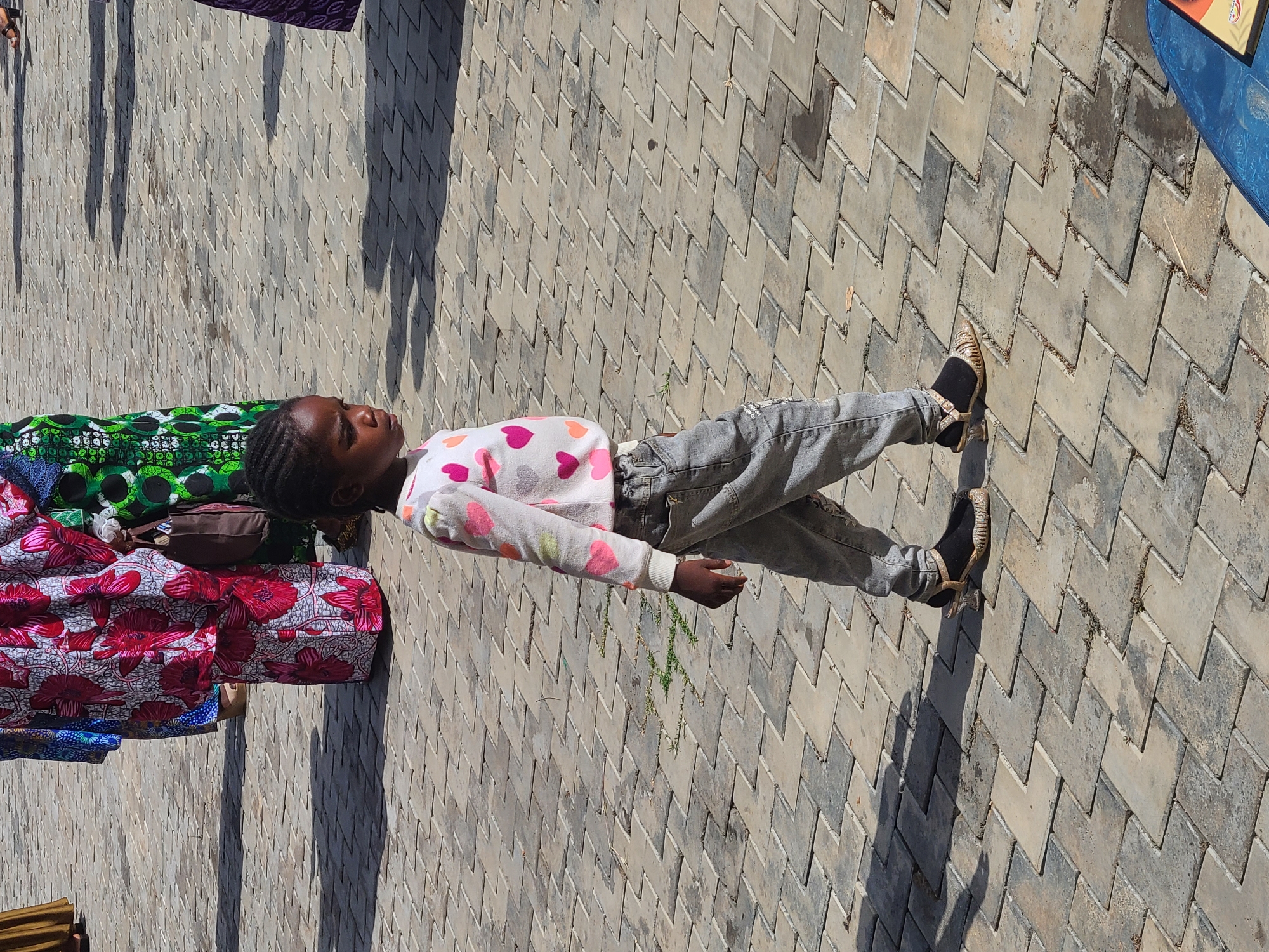 A girl lies on a gray, zig-zag brick pavement in Unknown, Unknown. She wears gray jeans, a light sweater with pink hearts, and patterned shoes. She rests her head on a stack of colorful fabrics. The pavement has patches of discoloration and some sparse vegetation.