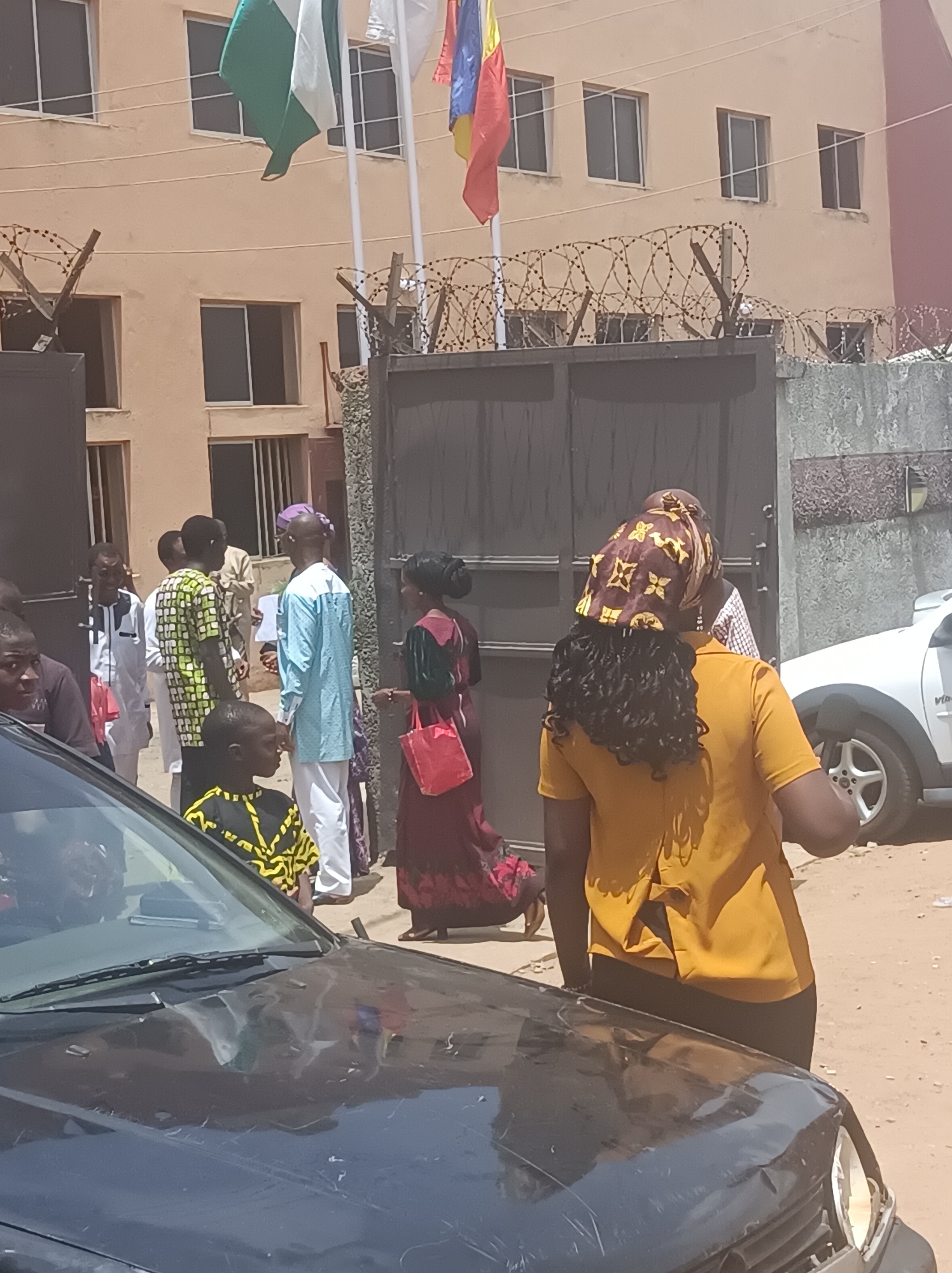 The image shows a street scene in Dadi Riba, Nigeria. Several people are gathered near a gate topped with barbed wire, outside of a building that is flying the flags of Nigeria, a white flag, and another flag that is partly obscured. Individuals are diverse in dress, some wearing traditional African clothing. There are cars nearby including the hood of a black car and a white SUV. The setting appears to be a sunny day.