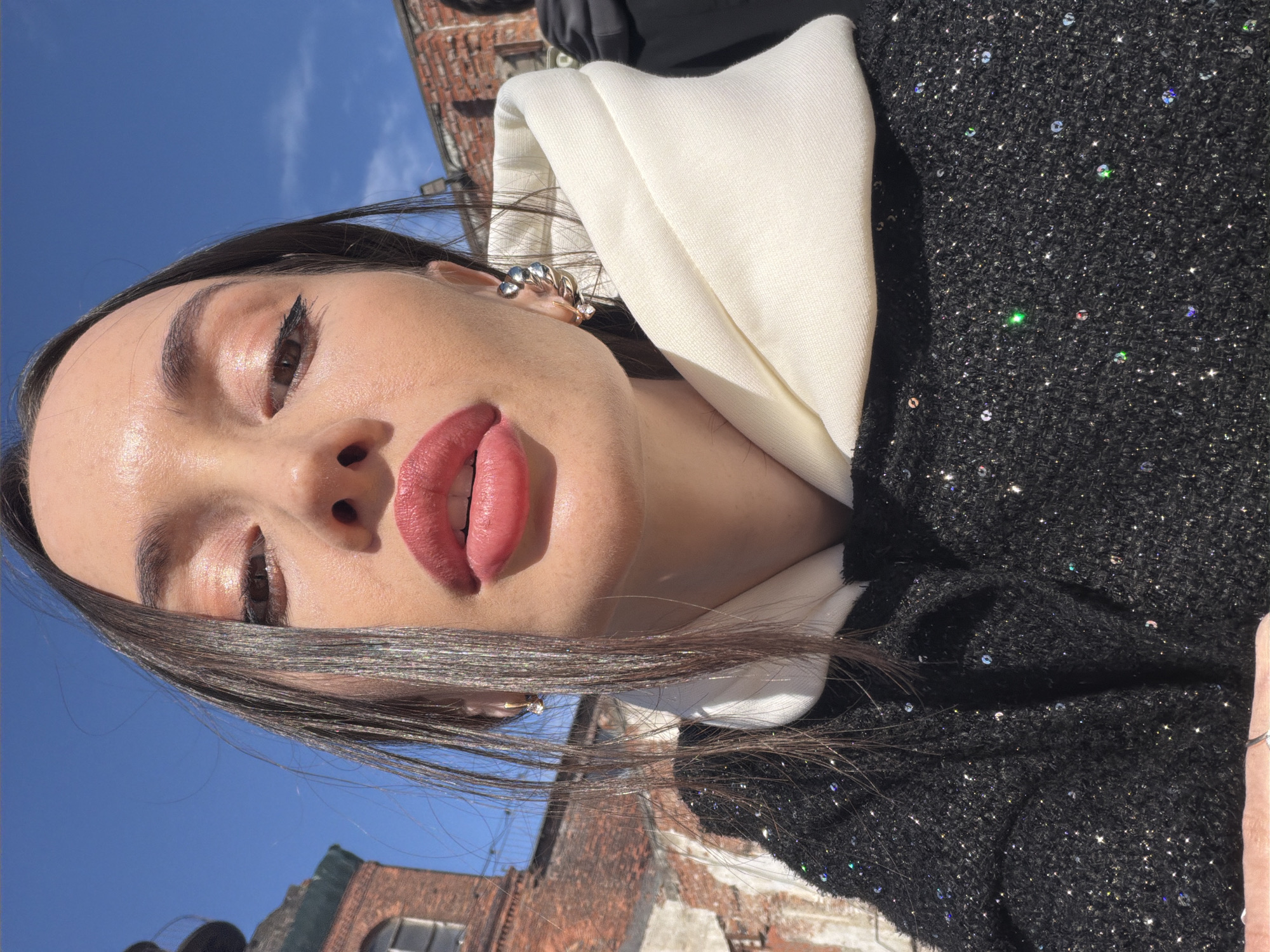 A close-up selfie of a woman is set against a partly cloudy blue sky and brick buildings. The woman is wearing a black, sparkly, tweed-like jacket with a large, white collar. She has dark hair, defined eyebrows, and is wearing eye makeup, with a dark liner. Her lips are painted with a red or pinkish-red color. She has silver earrings.
The buildings in the background appear to be brick and have architectural details, including windows. Based on the architectural style of the building in the background, it is likely that the location is Saint Petersburg, Russia.