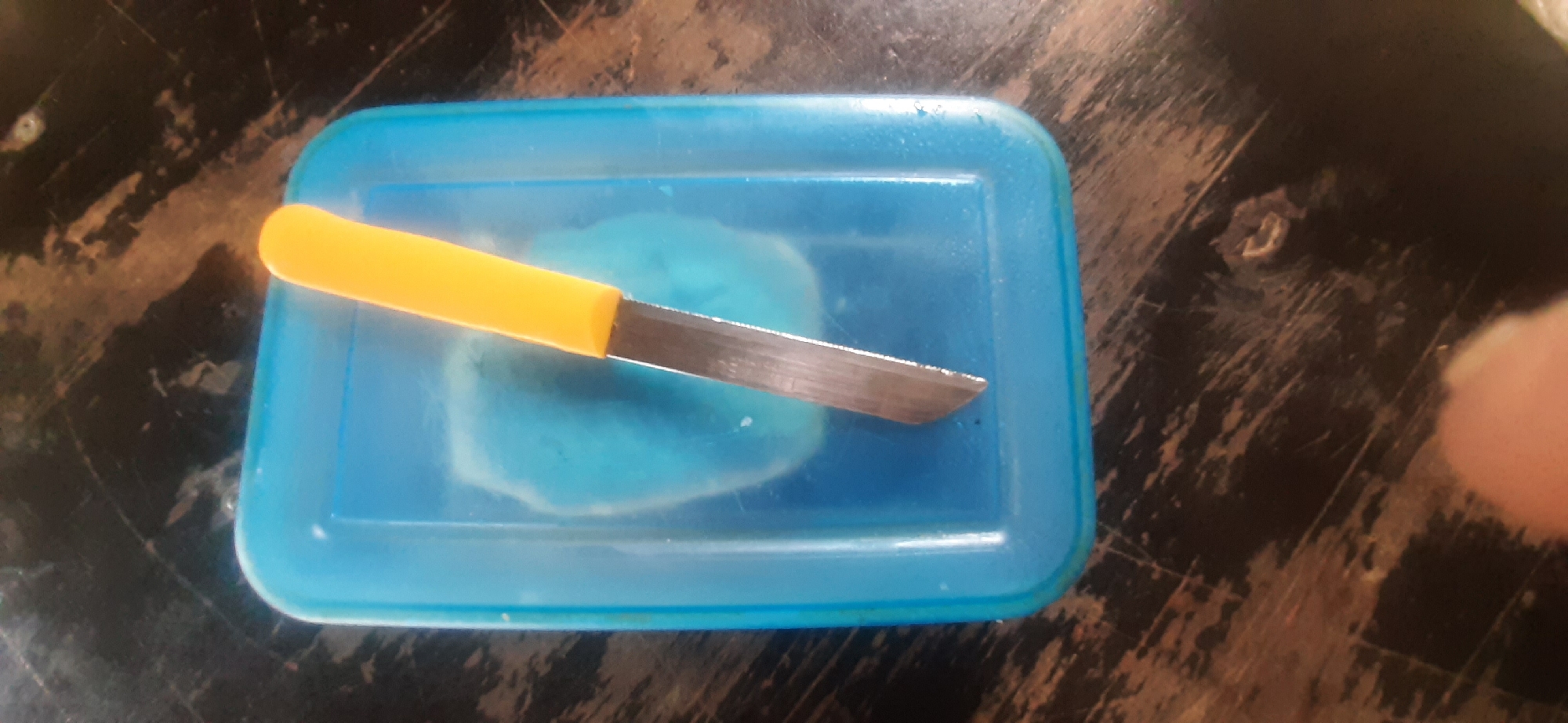 A knife with a yellow handle and a silver blade is positioned diagonally across a translucent blue container filled with a light blue substance. The container is resting on a dark, weathered surface that appears to be wood. The background is dark and textured, with visible scratches and imperfections on the surface. There are no people present in the image. The scene is captured from a slightly elevated, top-down perspective. The image does not provide any specific details or clues that would allow for its localization to Yessentukskaya, Russia.