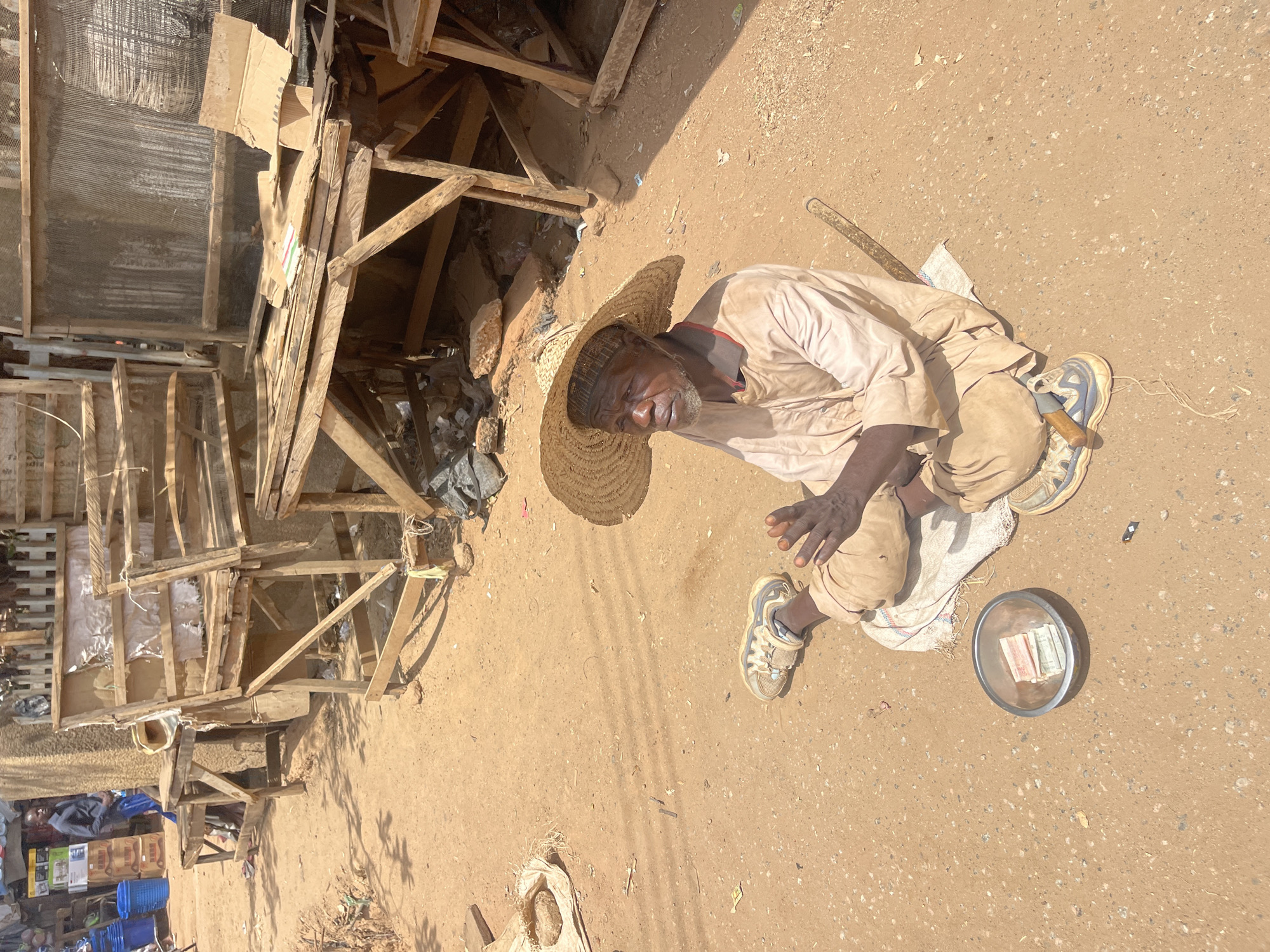 A man in Zaria, Nigeria, sits on the ground with a straw hat and wears a light brown outfit and sneakers. Next to him is a bowl containing folded currency. He is on the ground next to a wooden structure or stall with various objects behind it. The ground appears to be dry and dusty.