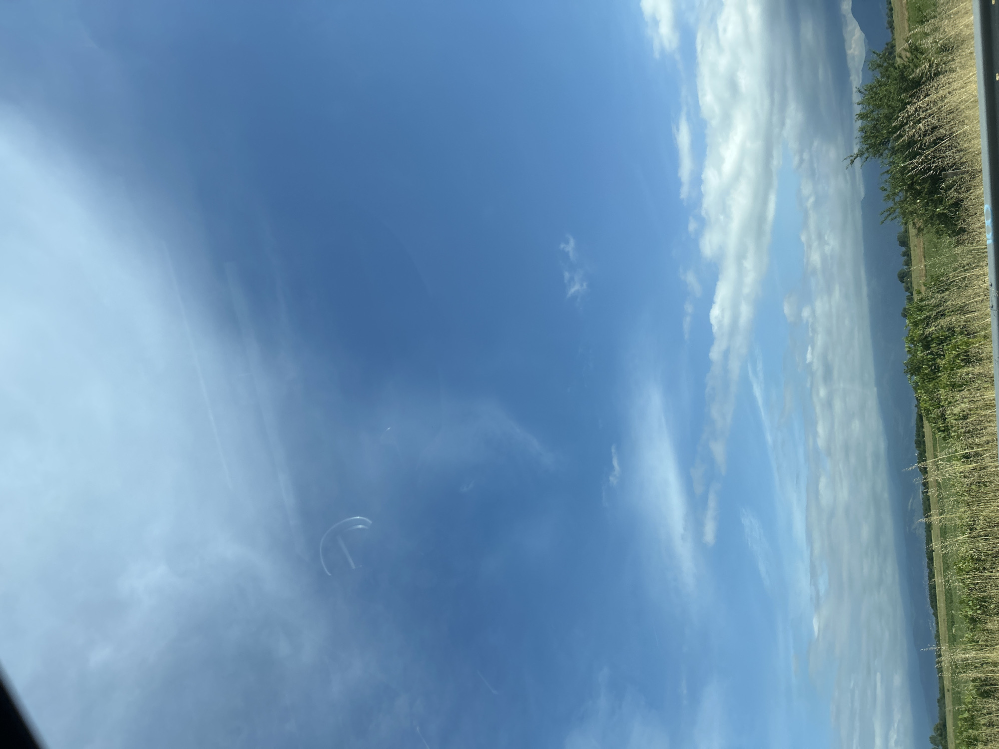 The image displays a wide outdoor landscape under a bright, sunny sky. The sky, which occupies the majority of the frame, is a vibrant blue, featuring scattered white clouds. Some clouds are thin and wispy, while others are more defined and cumulus-like, particularly towards the right side of the sky.
A clear horizon line separates the expansive sky from the land below. In the foreground, along the bottom of the image, there is a field of dry, tall grass or crops, suggesting an agricultural or rural environment. Beyond this field, a line of green trees and bushes forms a distinct treeline. In the far distance, subtle, hazy outlines of mountains or hills are visible against the lighter horizon.
The photograph appears to have been taken through a window, likely from a moving vehicle, indicated by reflections and glare on the glass. A noticeable semi-circular reflection is present in the upper left portion of the sky. The weather is clear and bright, consistent with daytime. There are no people or discernible text visible. The scene captures the rural and natural landscape, potentially indicative of the area around Malo Konare, Bulgaria, with its agricultural land and distant mountainous terrain.