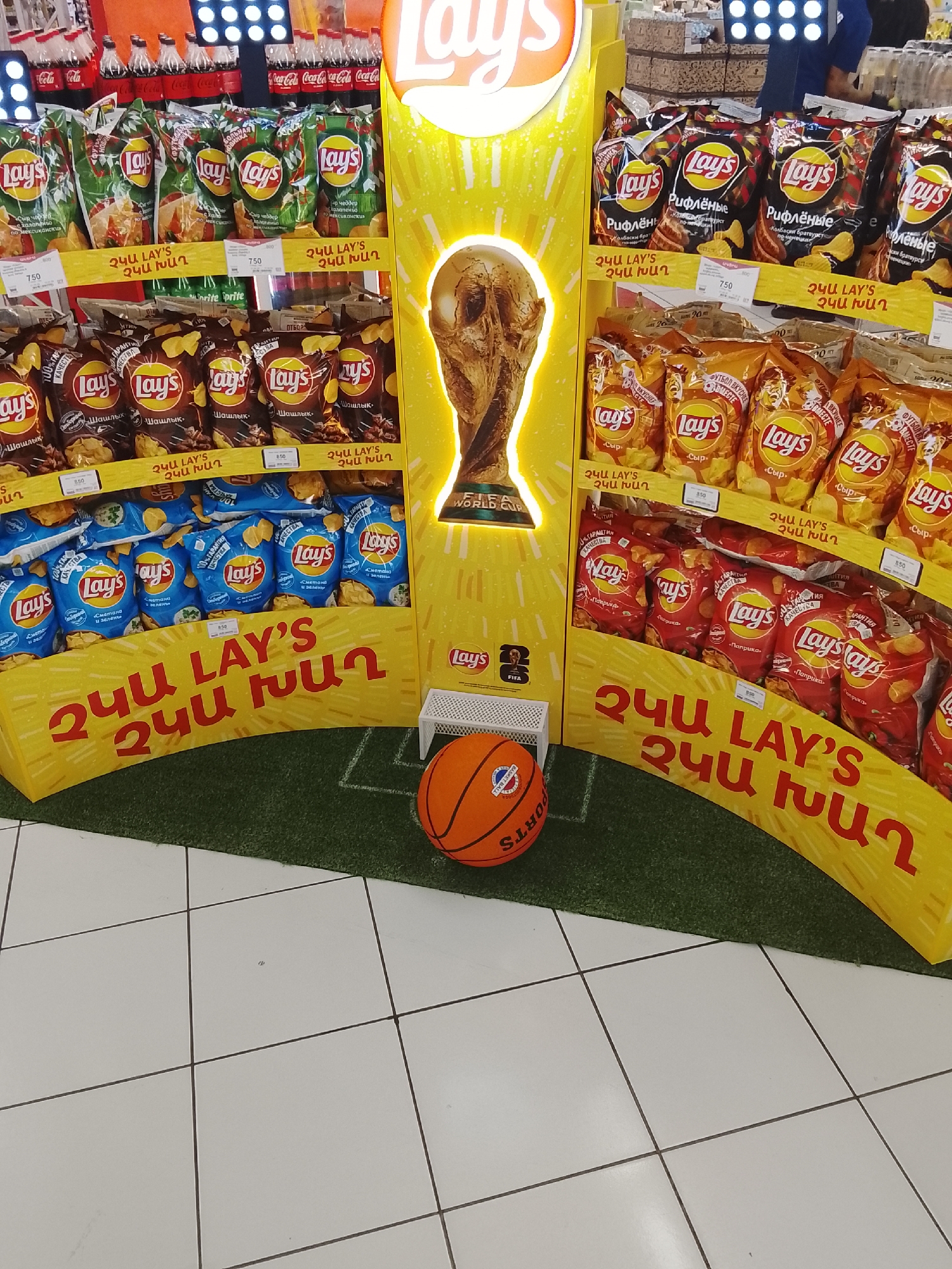 An eye-level, wide shot captures a brightly lit grocery store display. The focal point is a tiered stand showcasing various flavors of Lay's potato chips, arranged in colorful bags. The display is adorned with a large, illuminated replica of the FIFA World Cup trophy. At the base of the display, a miniature soccer field is simulated with green turf, complete with white lines. A basketball sits on the turf, hinting at a sports theme. The background shows rows of soda bottles and other merchandise, along with aisle lighting. The flooring appears to be white tile. The overall impression is a promotional setup designed to attract shoppers with a combination of popular snack brands and sports imagery.