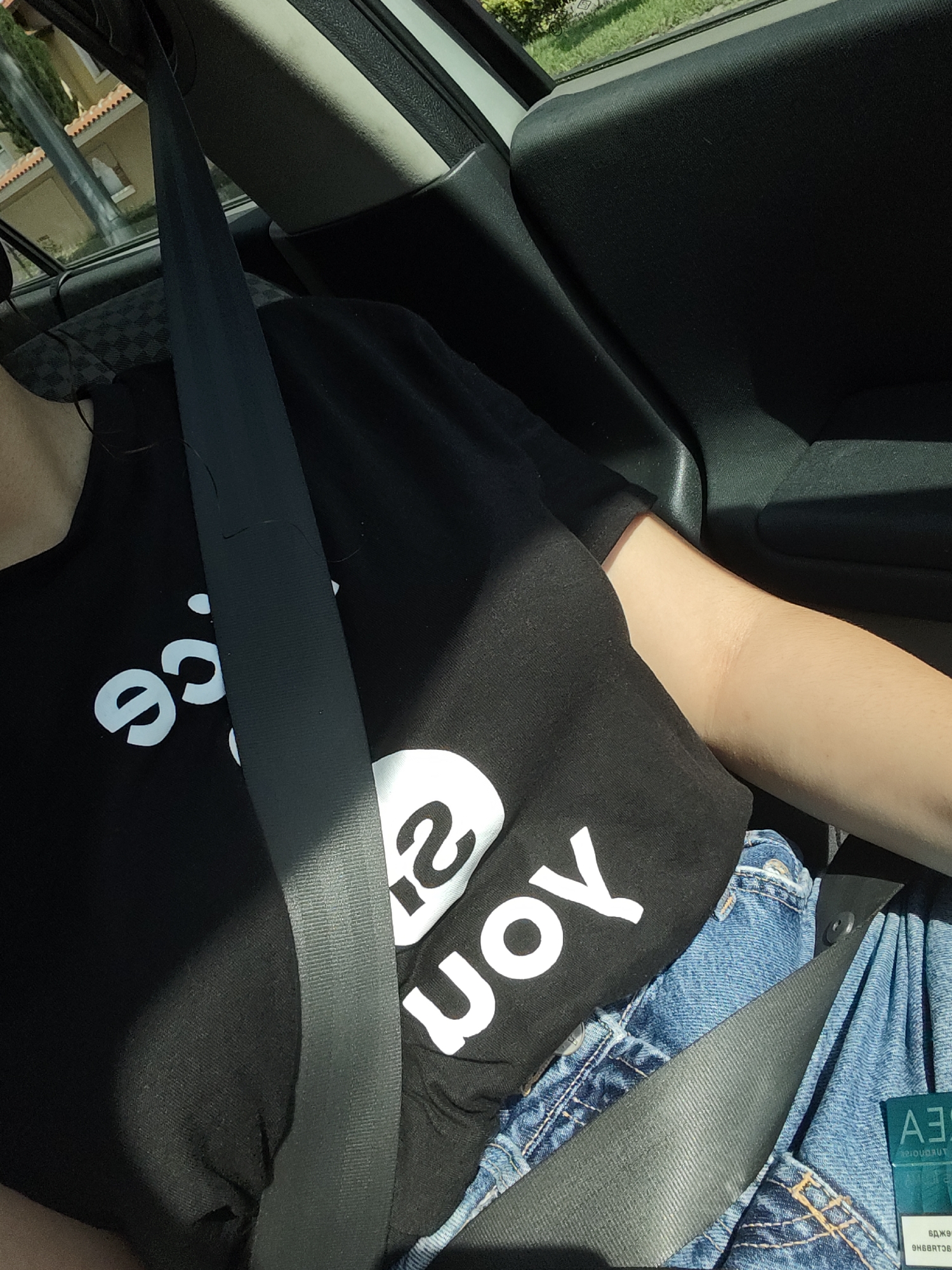 A person is seated in a car, wearing a black t-shirt with white text and blue denim jeans. A grey seatbelt is fastened across their chest and lap. The person's arm, with visible light hair, is resting on their lap. The interior of the car is visible, including a black dashboard and a portion of the car door. Outside the car window, a building with an orange tiled roof and greenery can be seen. A small blue and white card with text is partially visible in the lower right corner of the image.