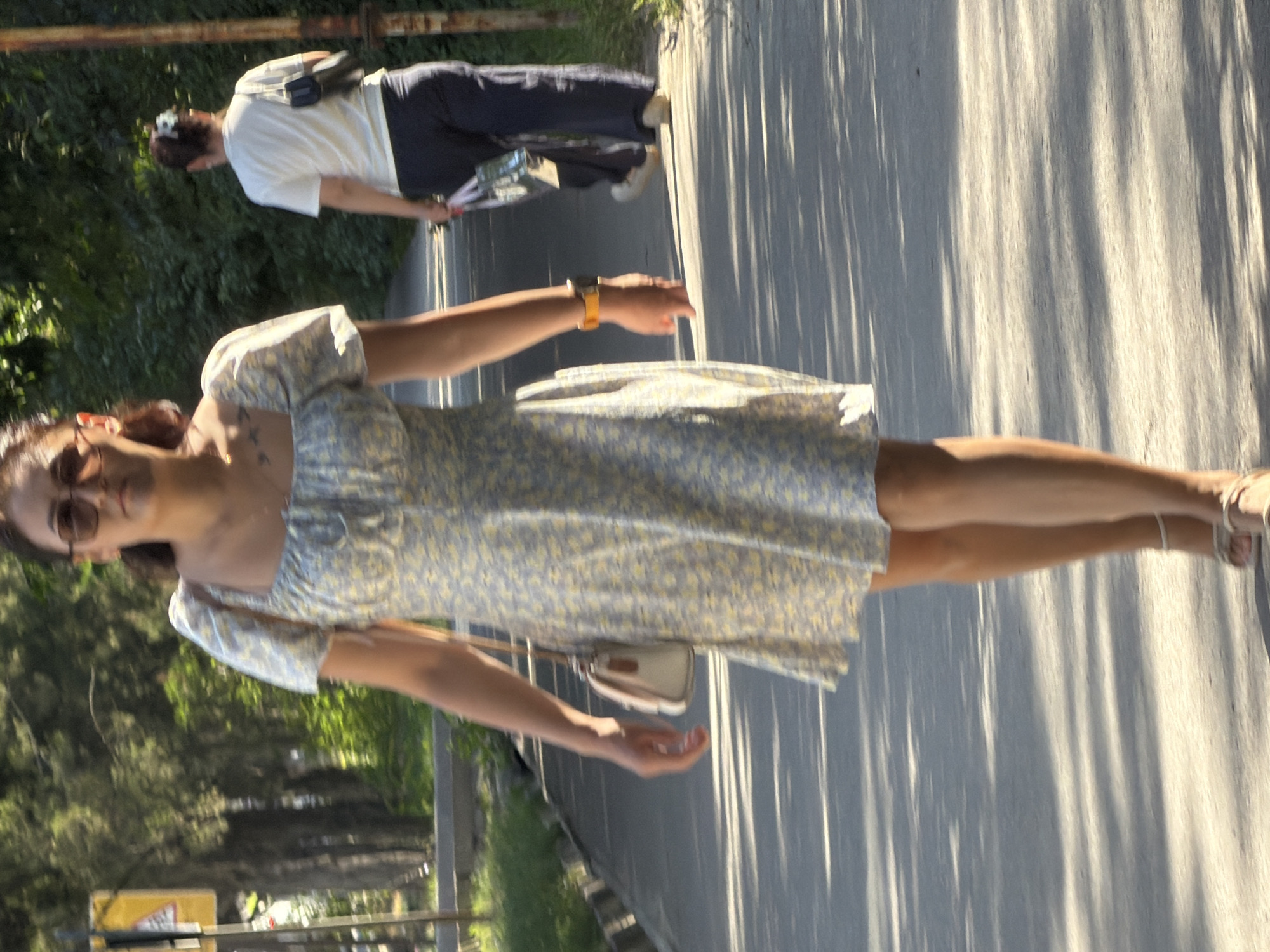 An outdoor, eye-level medium shot captures two individuals walking on a paved pathway under dappled sunlight. In the foreground, a woman walks towards the camera. She has her hair pulled back and is wearing sunglasses, a light-colored floral dress, and white sandals. A small, light-colored purse hangs from her left shoulder. She is also wearing an orange watch on her left wrist. Her right arm is slightly raised with her hand open.
In the background, another person, viewed from behind, walks away from the camera. They are wearing a white t-shirt, dark pants, and light-colored shoes. This individual carries a dark bag over their right shoulder and holds a few items in their left hand.
The pathway is bordered by a curb and grass on the left, and trees and foliage on the right. The background shows dense greenery, indicating an outdoor environment. The sunlight casts distinct shadows across the pathway, suggesting it is daytime. A yellow triangular sign is visible on the left side of the frame, and a rusty pole stands on the right.