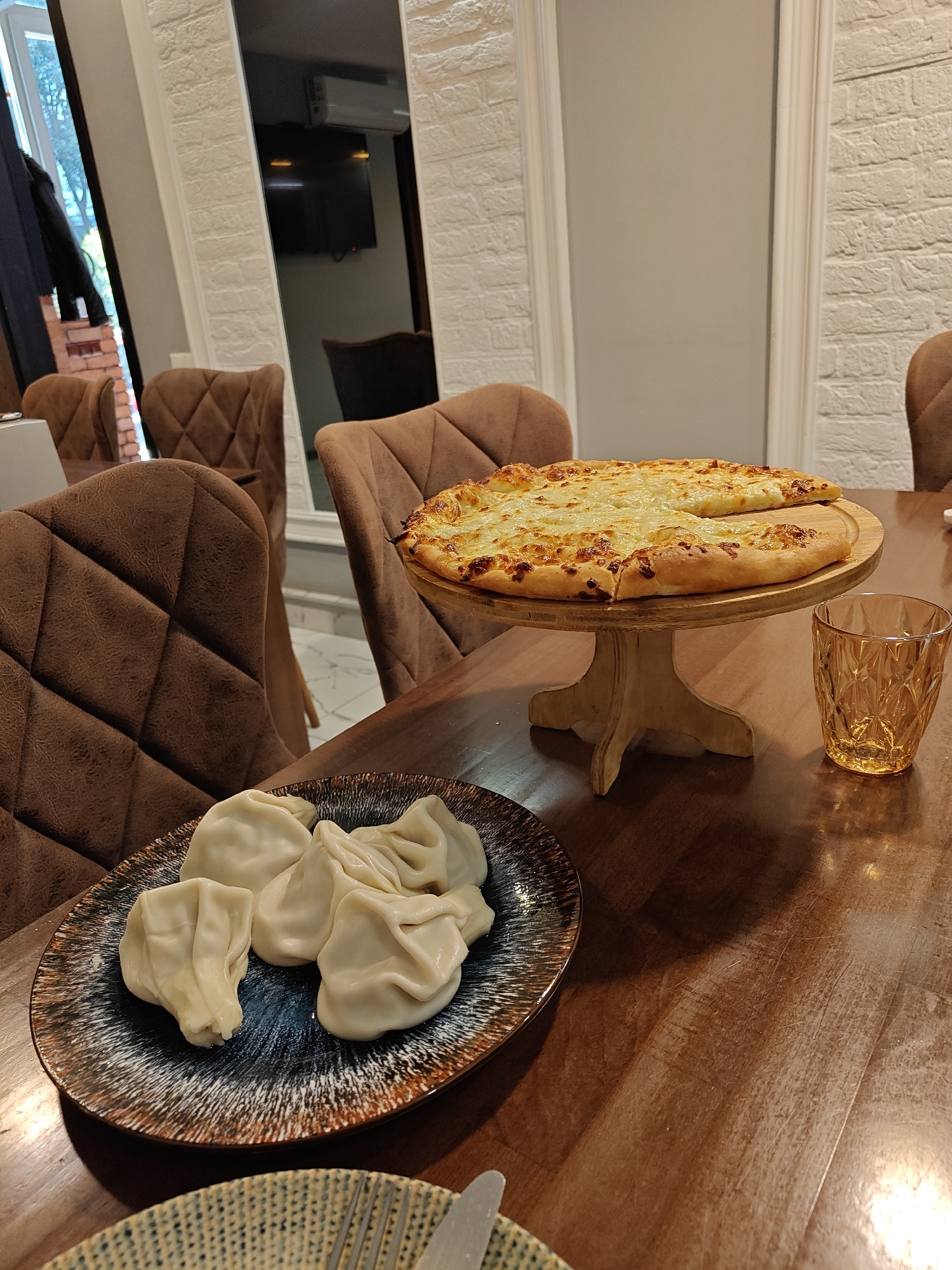 The image depicts a table setting in what appears to be a restaurant in Batumi, Georgia. Central to the image are two traditional Georgian dishes: khinkali and khachapuri. Khinkali, large Georgian dumplings, are arranged on a blue and brown patterned plate. Adjacent to the plate, a golden glass sits on the dark brown wooden table. In the background, a circular wooden stand holds a partially eaten khachapuri, a Georgian cheese bread. Brown upholstered chairs surround the table. The background shows a mirror, giving a partial view of other parts of the room. The setting indicates an indoor dining area. The presence of these particular dishes strongly points to the location being in Georgia, likely Batumi due to the style of restaurant.