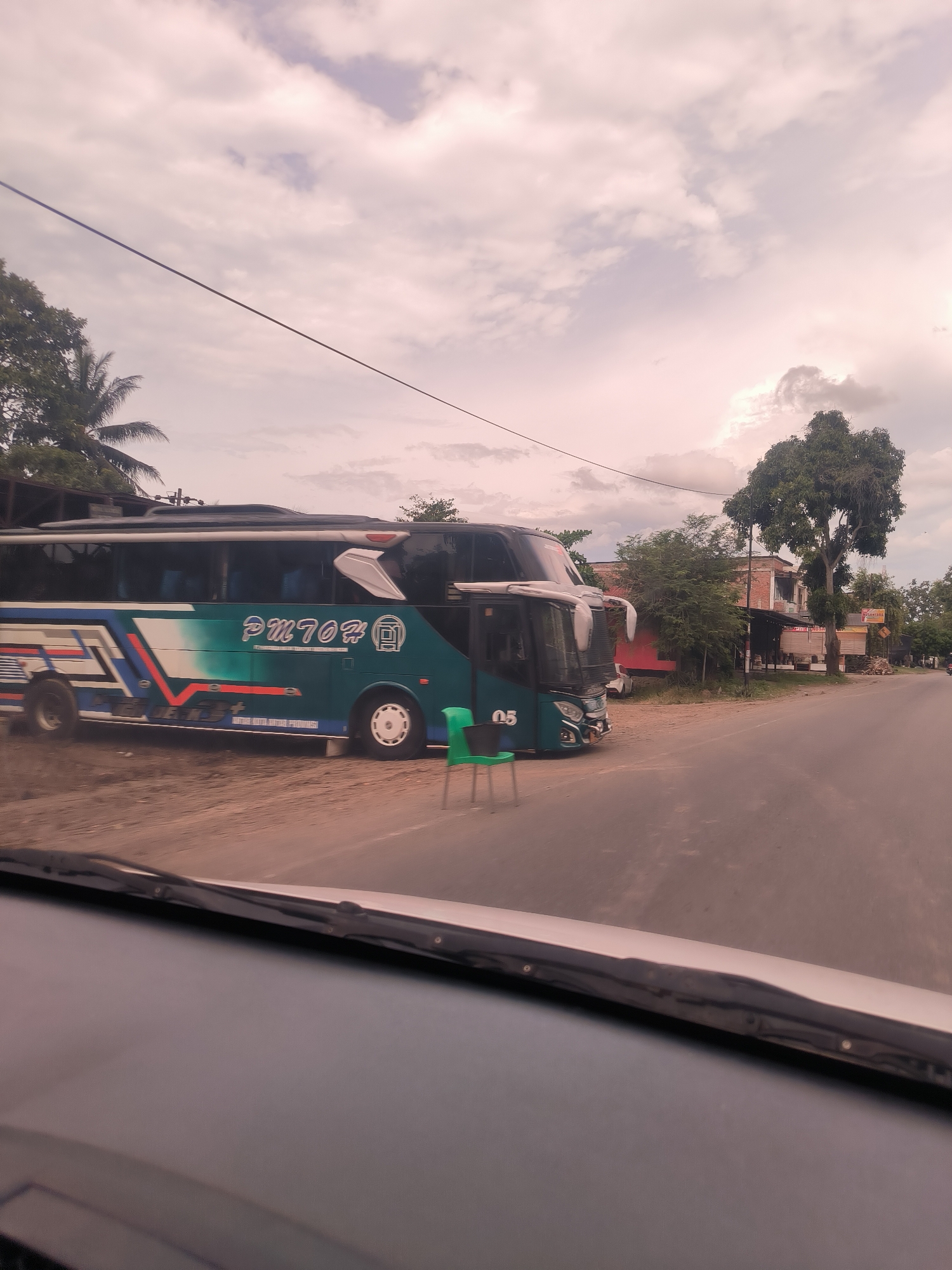The image depicts a static scene in Reuhattuha, Indonesia, viewed from inside a vehicle, with the dashboard and windshield wipers partially visible in the foreground.
The primary subject is a large, double-decker passenger bus, colored teal and white with blue and red stripe accents. It is parked on a dirt shoulder beside an asphalt road. The bus prominently displays "PM 702 #" on its side, along with the number "05" near its front wheel. Smaller text, partially visible below "PM 702 #", appears to read "PUTRA NAGA ATJA PERMAI". A bright green plastic chair is positioned in front of the bus, near the road's edge.
The scene is set under a cloudy sky, with soft, diffused light suggesting either early morning or late afternoon. To the left of the bus, dense tropical foliage, including palm trees, is visible. To the right, along the road, more trees and several buildings, including a red-bricked structure, line the street. A sign on one of the distant buildings in the background might read "KECAP NAGA" or a similar phrase, though it is not entirely clear. No people are visible in the image. The overall atmosphere is quiet.