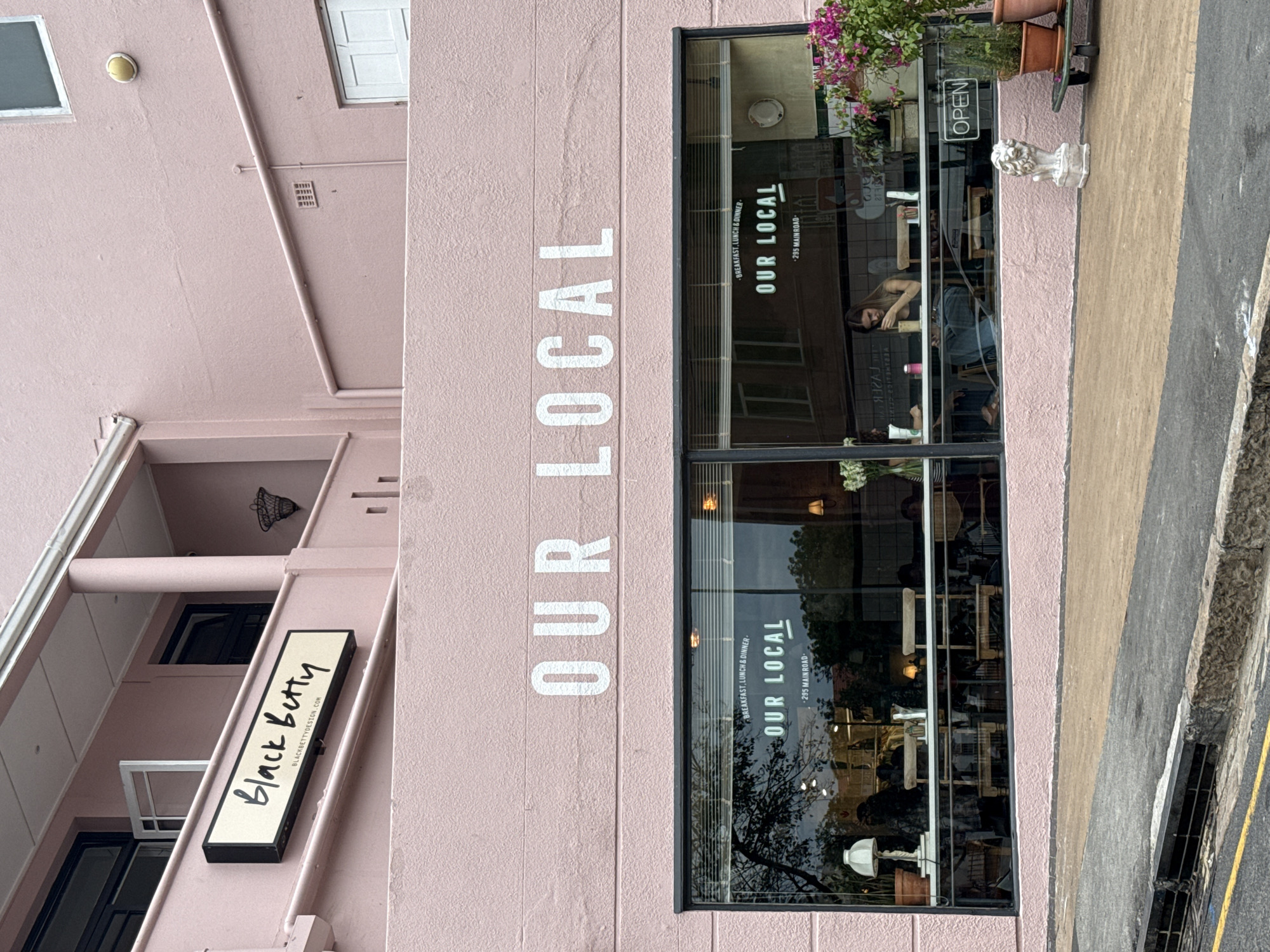 The image shows a pink building with two storefronts, "Our Local" and "Black Betty", and is presumably located at or near 295 Main Road. "Our Local" has a sign in white lettering on the facade, and the storefront windows display the words "OUR LOCAL", "BREAKFAST, LUNCH & DINNER" and "-295 MAINROAD-". The windows also reflect the outside environment, as well as displaying seated customers inside. "Black Betty", on the left side of the image, has a black and white sign indicating the business's name. The location is likely in Cape Town, South Africa, based on the address. The overall scene appears to be a street view, showing a business district with a restaurant and design store.