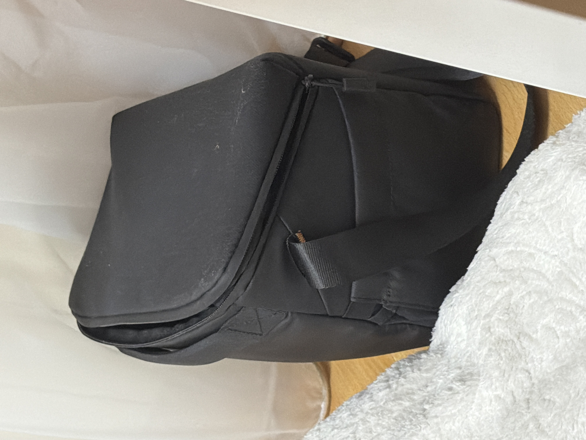 A black bag sits on a wooden surface. The bag is made of a matte material and has a zipper closure. A white towel is partially visible in the foreground, to the left of the bag. A white curtain is visible in the background. The scene appears to be indoors.