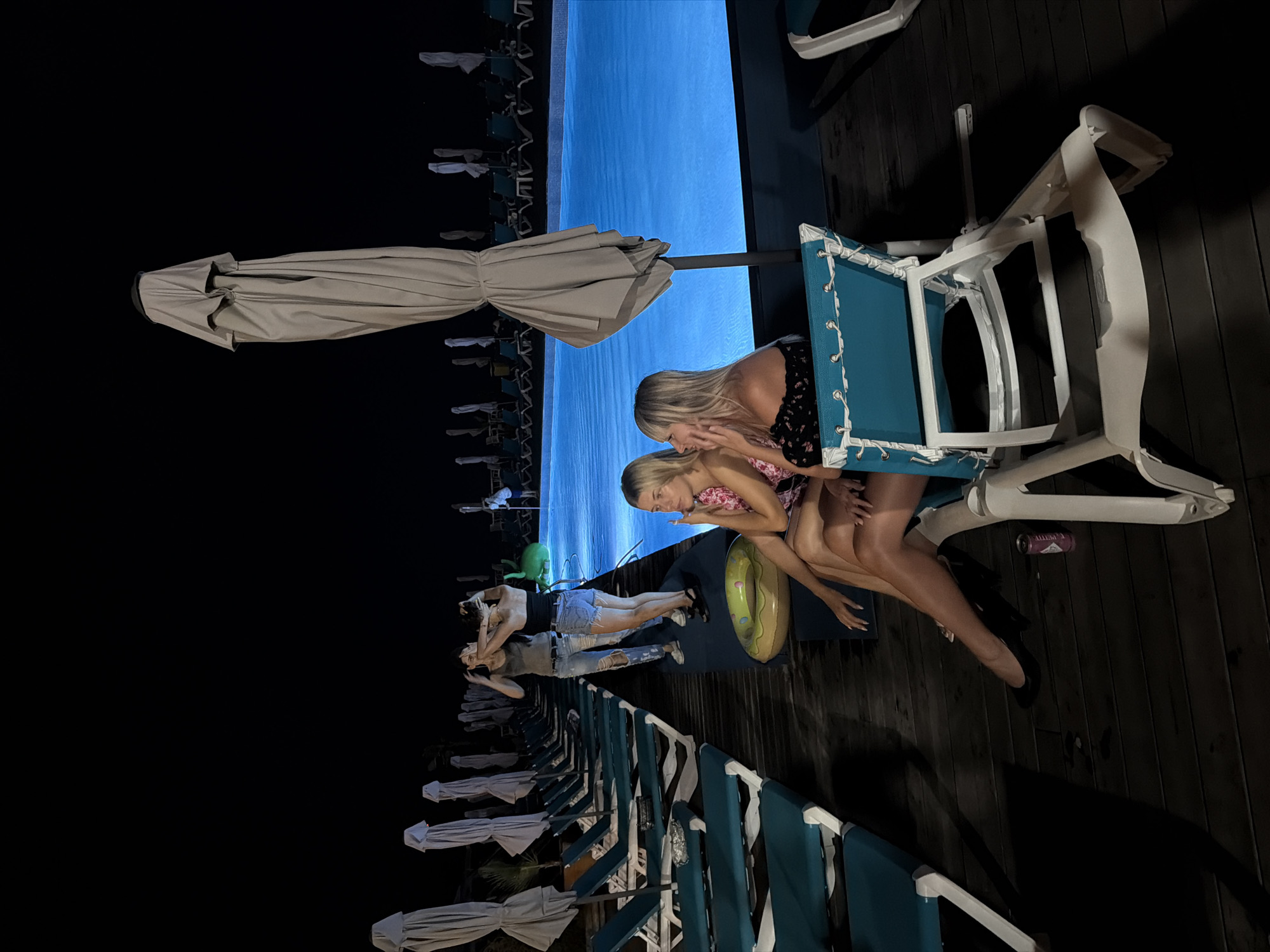 At night, two women are seated on lounge chairs next to a bright blue swimming pool. The woman on the left, with blonde hair, wears a floral-patterned top and a black skirt. The woman on the right, also blonde, is dressed in a black off-the-shoulder top and a black skirt. They are positioned on a wooden deck, with several other empty lounge chairs and closed umbrellas lining the area. In the background, three other women are standing near the edge of the pool. One woman, wearing a grey tank top and ripped jeans, has her hands on her head. Another woman, in a black top and denim shorts, also has her hands near her head. A third woman, in a green outfit, is visible further away. The pool is illuminated by underwater lights, casting a vibrant blue glow. The scene appears to be at a resort or hotel in Sirius, Russia, given the provided location.