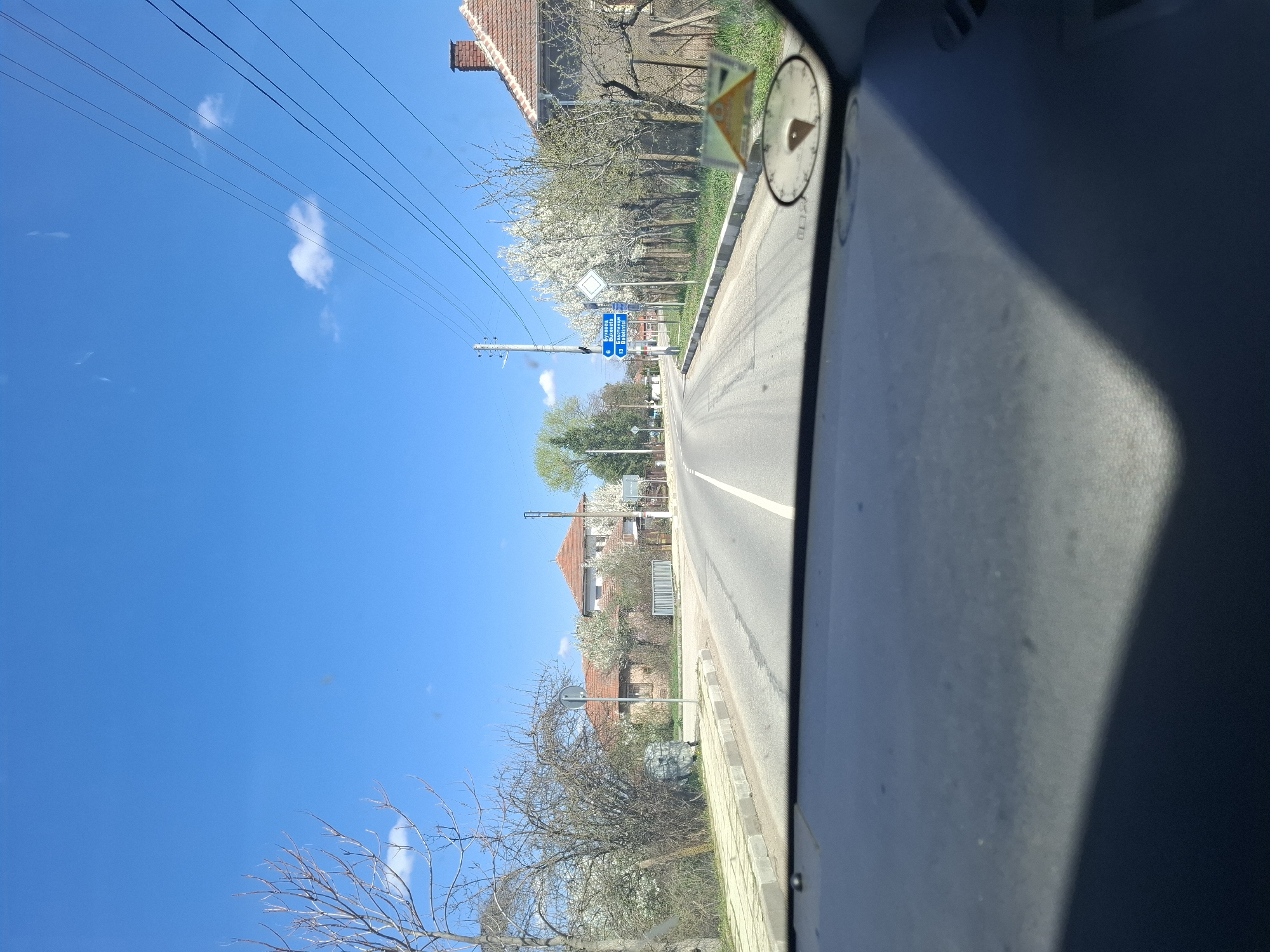 Captured from inside a vehicle moving along a road, the image presents a daytime scene in Bukovets, Bulgaria. The asphalt road stretches forward with a solid white lane marking its center. Alongside the road, there is a sidewalk. The environment includes residential buildings with red-tiled roofs and various trees, some in bloom. Utility poles with overhead power lines run along the road. Street signs with blue backgrounds and white lettering are visible. A sticker is present on the vehicle's dashboard. The sky is clear blue with a few clouds.