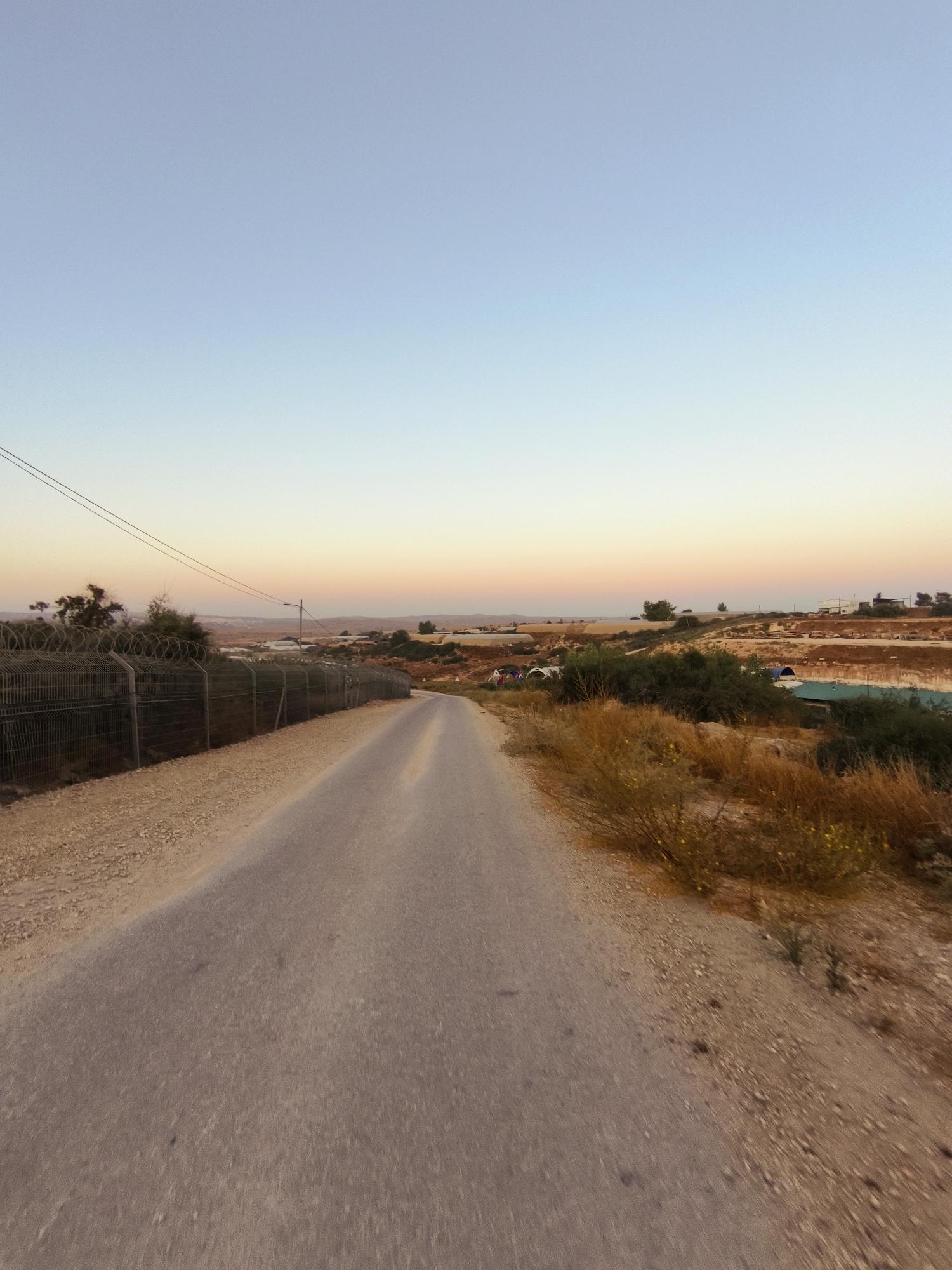 An aerial drone shot captures a paved road stretching into the distance, flanked by dry brush and a barbed-wire fence on the left. The road winds through a landscape dotted with arid vegetation, some low buildings, and what appear to be agricultural structures or greenhouses. The sky transitions from a pale blue at the top to soft hues of pink and orange near the horizon, suggesting either sunrise or sunset. The scene is devoid of human presence and activity. The location is the Lachish Regional Council, Israel.