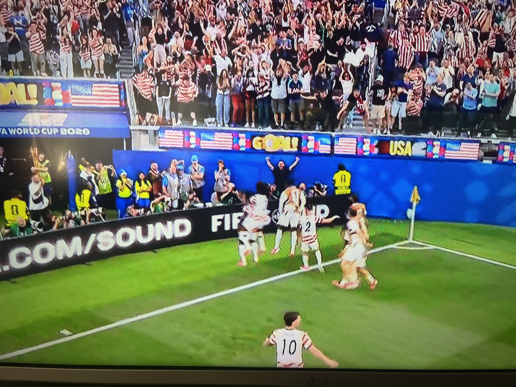 The image shows a soccer game in a stadium, likely during a World Cup event, given the "FIFA WORLD CUP 2020" banner visible. The field is green, with white markings indicating the boundaries and penalty areas. In the foreground, a player wearing a white jersey with the number 10 is on the field. In the background, several players in white and red striped jerseys are celebrating, with one player on the ground. A large crowd is visible in the stands, many wearing American flag-themed attire. The stadium appears to be outdoors, with a blue backdrop behind the crowd and banners. The overall atmosphere suggests a moment of excitement and victory for the team in white and red.