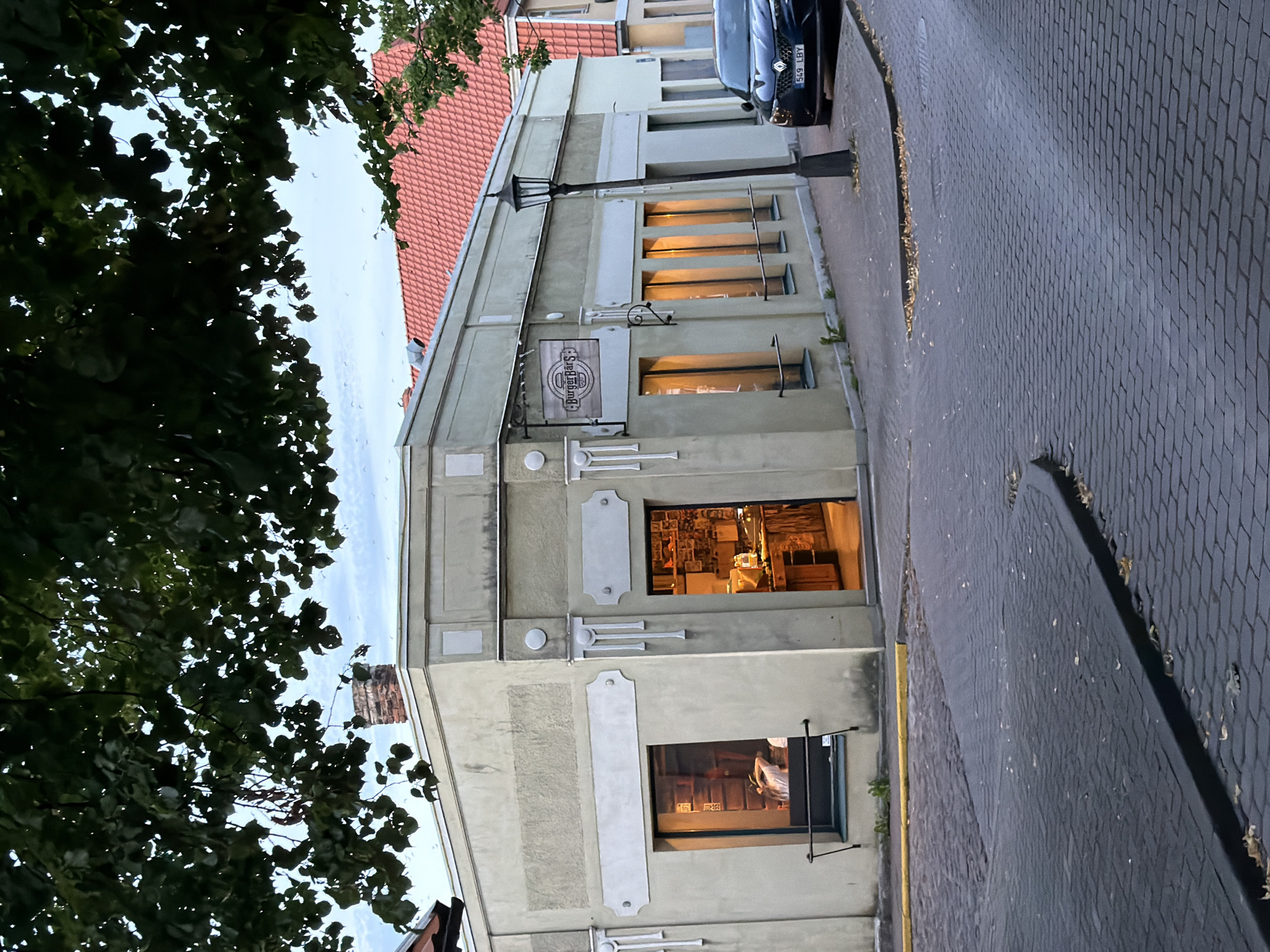 The image depicts a corner building in Ventspils, Latvia, adorned with a sign that reads "BurgerPārs". The building, painted in a light yellow-beige color, features decorative elements around its windows and entrance. The entrance, illuminated by warm interior lights, reveals a glimpse of the restaurant's interior with shelves and what appears to be a counter. To the left of the entrance, a large window displays shelves and a person with their back to the camera. The building is situated on a cobblestone street, with a black Renault car parked to its right. A lamppost stands in front of the restaurant. Overhanging trees with dark green foliage partially obscure the sky, which is a soft blue with a hint of white clouds. The overall scene suggests a quiet street in Ventspils, likely in the evening or early morning, with the restaurant warmly lit, indicating it is open or recently closed.