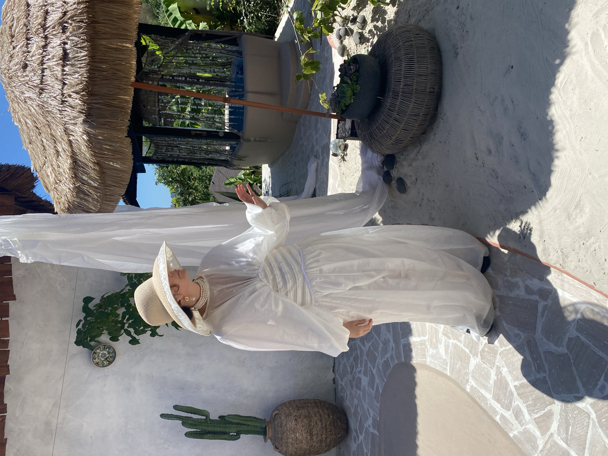 A full shot shows a woman in a white dress and a wide-brimmed hat standing in what appears to be an outdoor courtyard in Sochi, Russia. The woman is looking to her left with her right hand extended. She is wearing a pearl necklace and earrings. The woman is standing on a stone patio next to a white curtain. In the background, there is a thatched umbrella over a circular structure with blue water inside. There are also plants and trees surrounding the area. The sun is shining brightly, casting shadows on the ground.